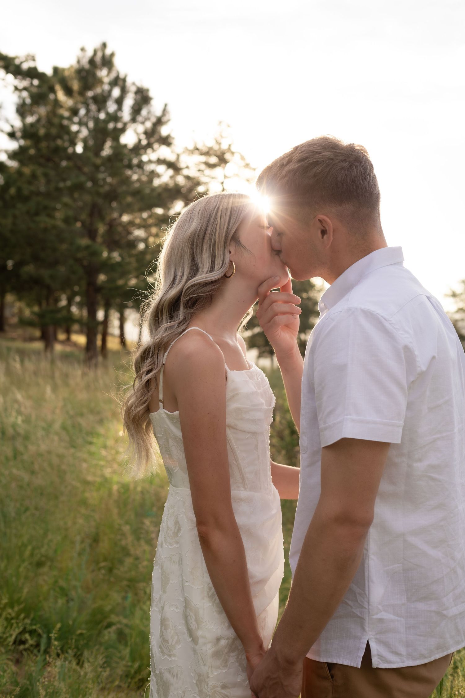 Couple kissing at golden hour in a sunlit meadow, woman in white dress holding hands with man.