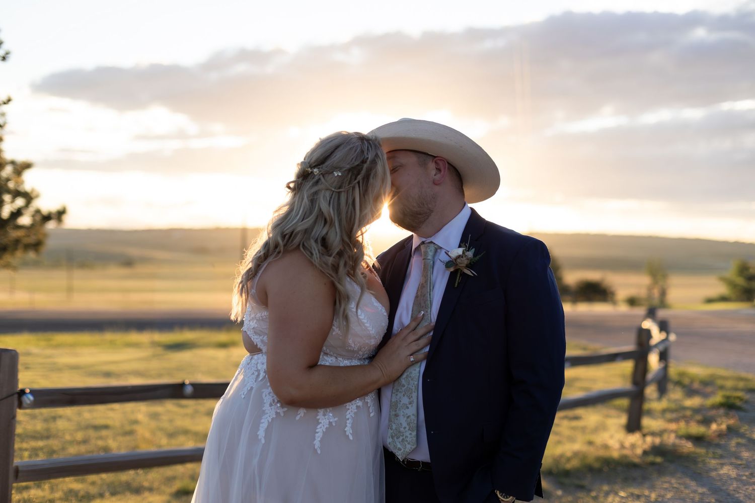 Bride and groom sharing a kiss at golden hour by a wooden fence in an open field during an outdoor country wedding.