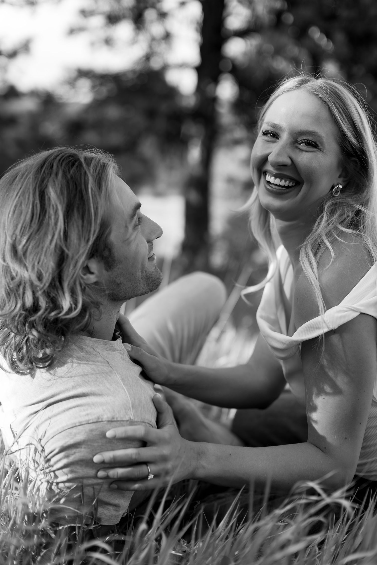 Black and white photo of a laughing woman embracing a man while sitting together in grass outdoors.