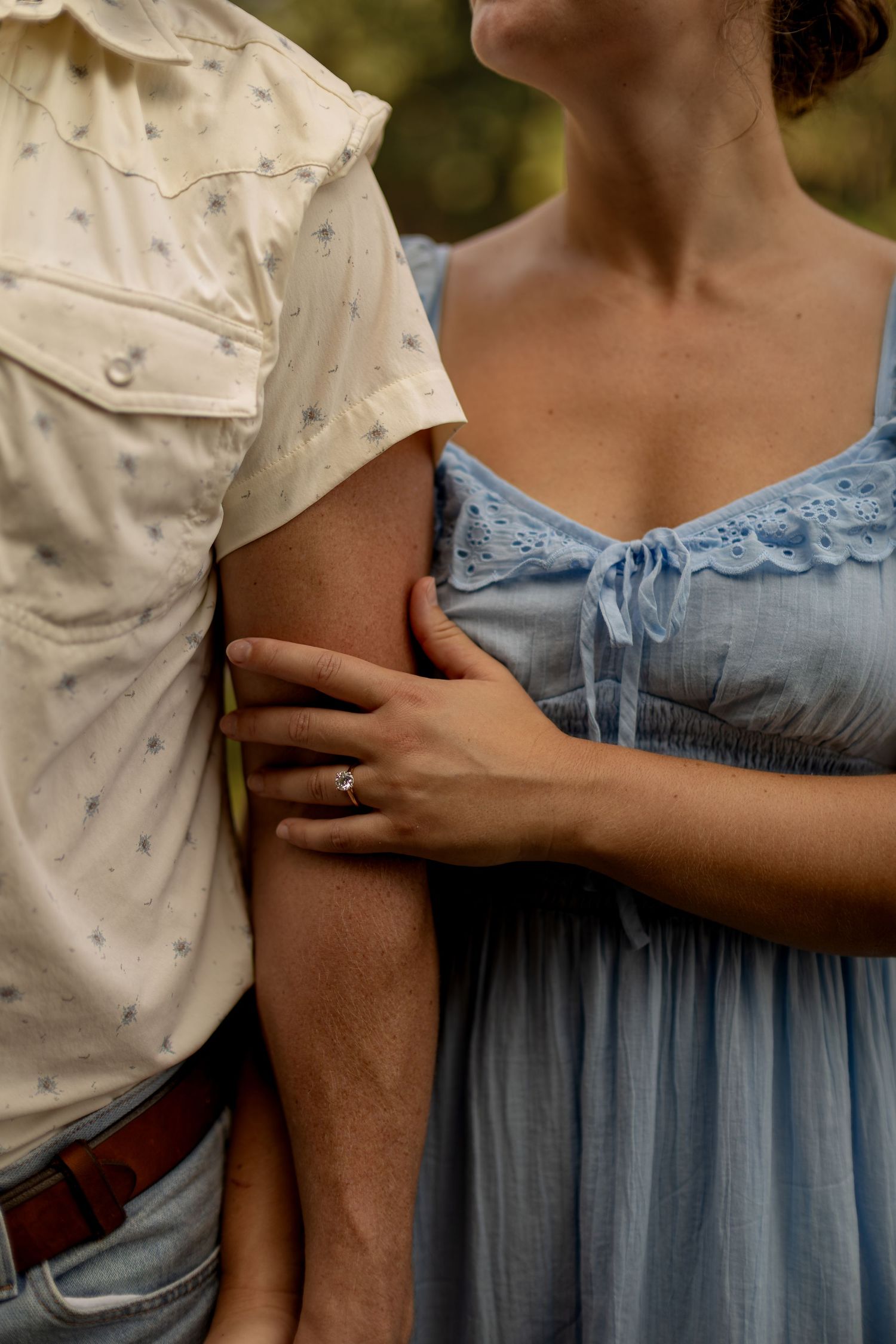 Close-up of couple standing together, woman in blue dress holding mans arm, wearing engagement ring.