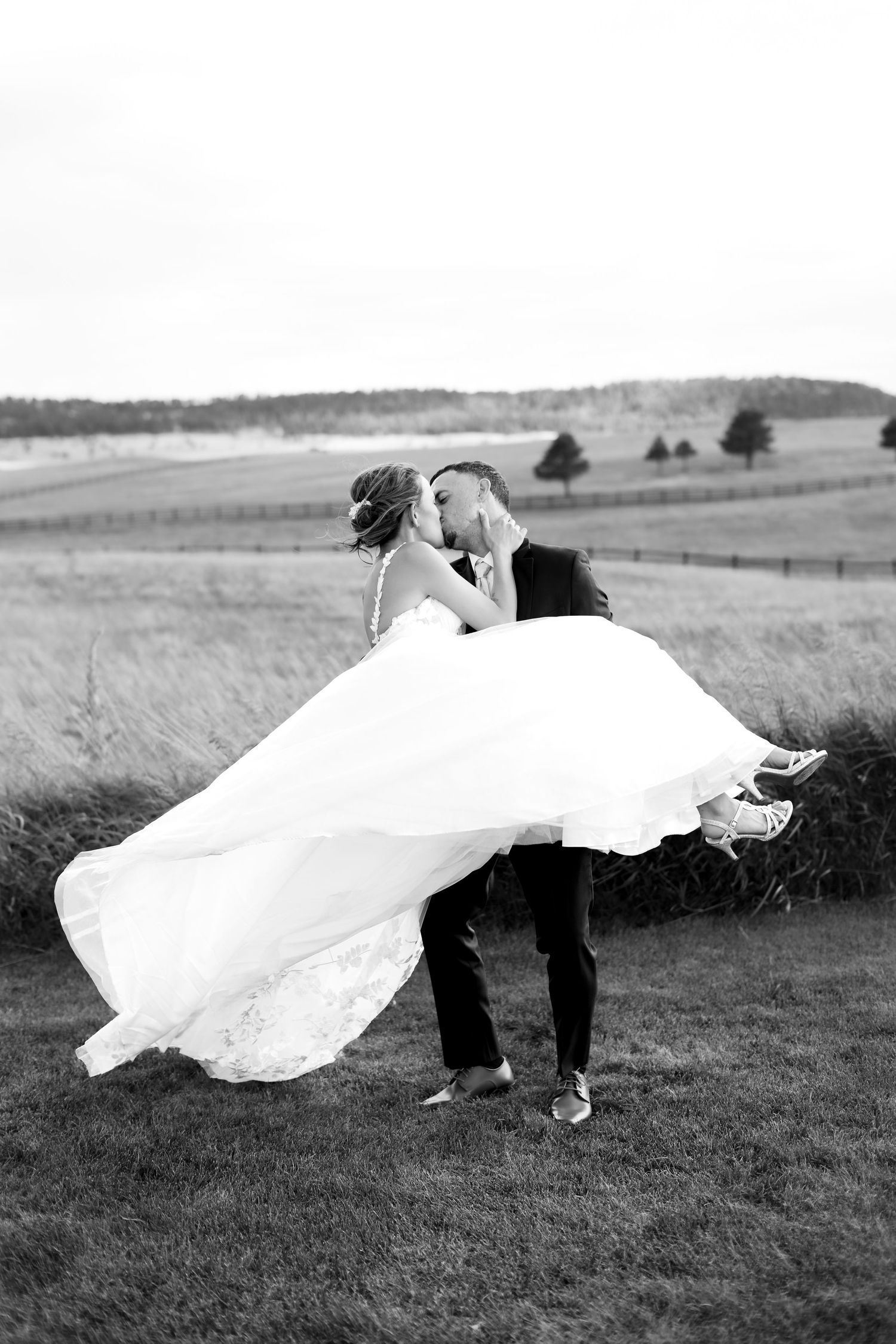Groom lifts and spins bride in a flowing white dress while kissing in a scenic outdoor field.
