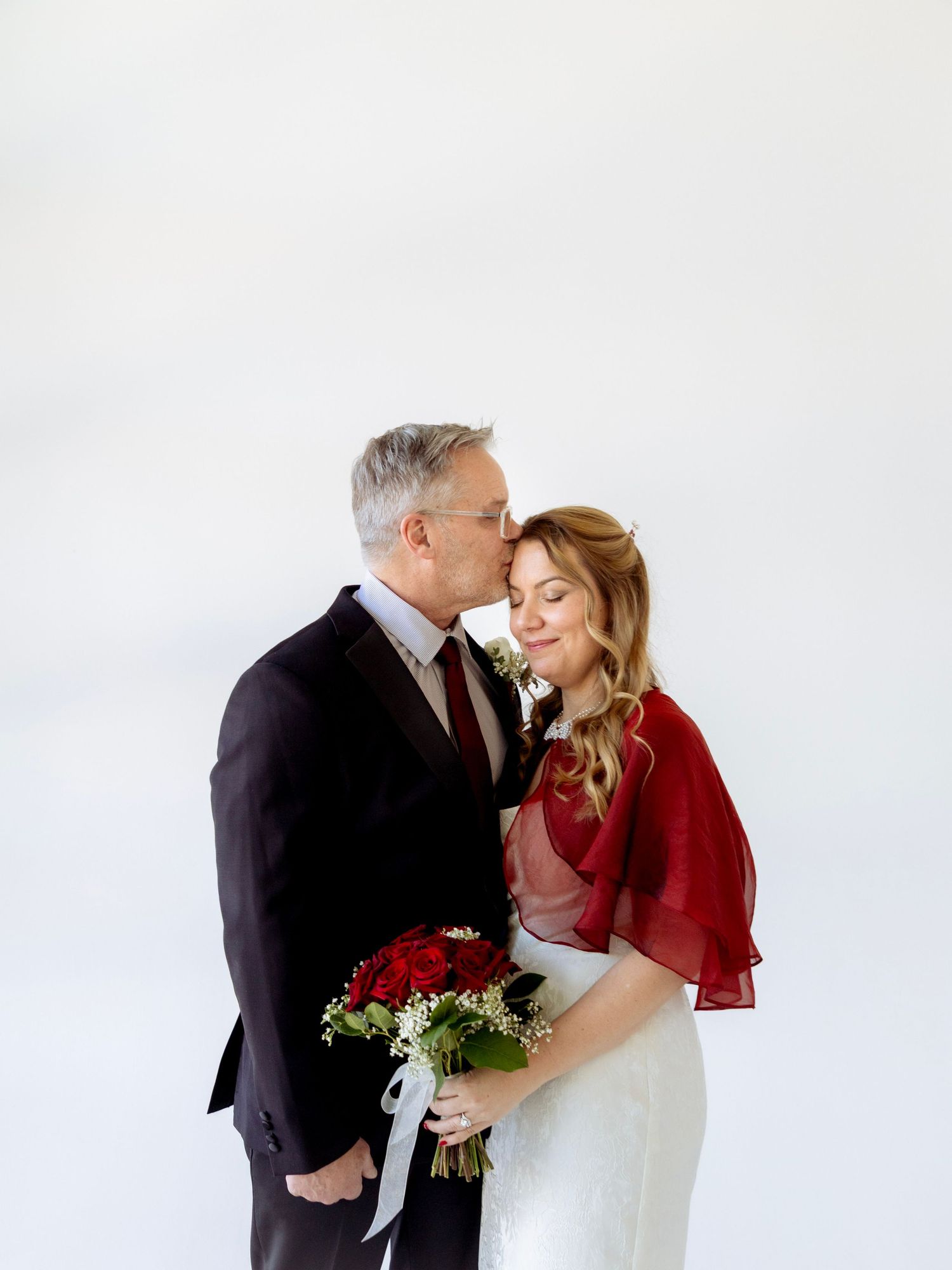 Elegant couple in wedding attire sharing a tender kiss, bride holding red roses bouquet, against white background.