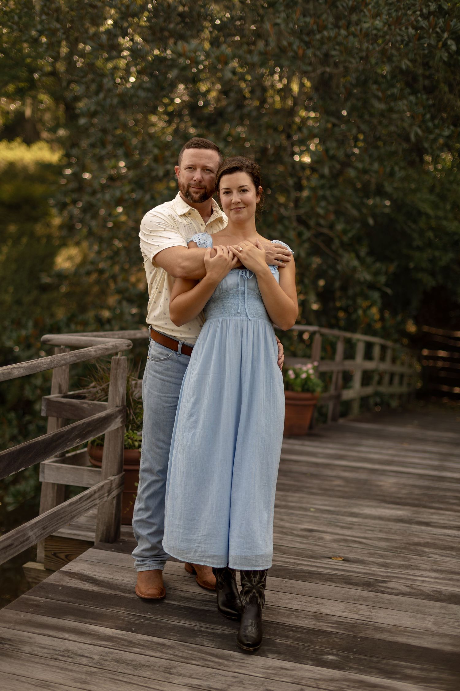 Couple embracing on a wooden bridge surrounded by lush green trees, woman in blue dress, man in white shirt and jeans.