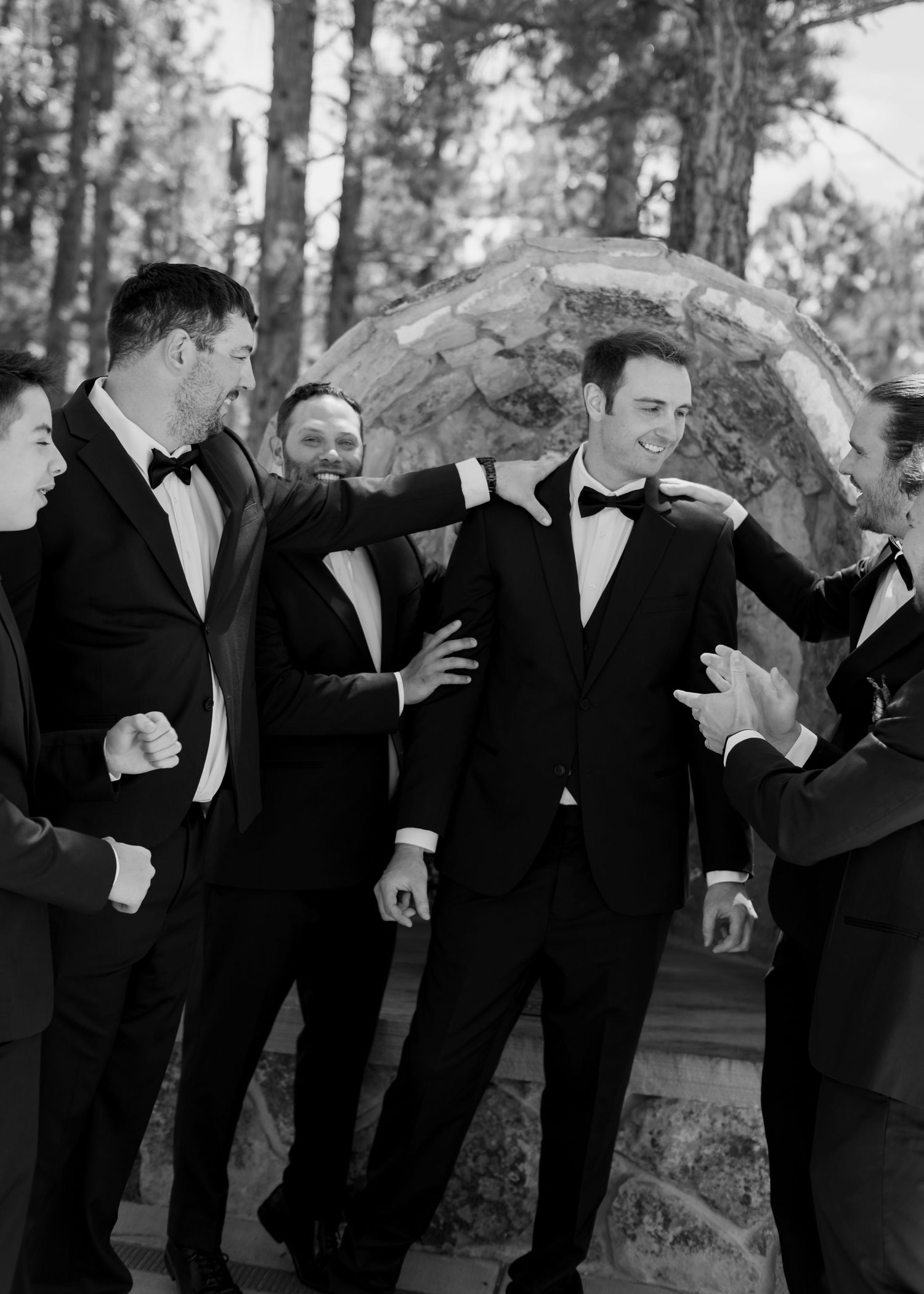 Black and white photo of groom surrounded by smiling groomsmen in tuxedos celebrating outdoors near trees.