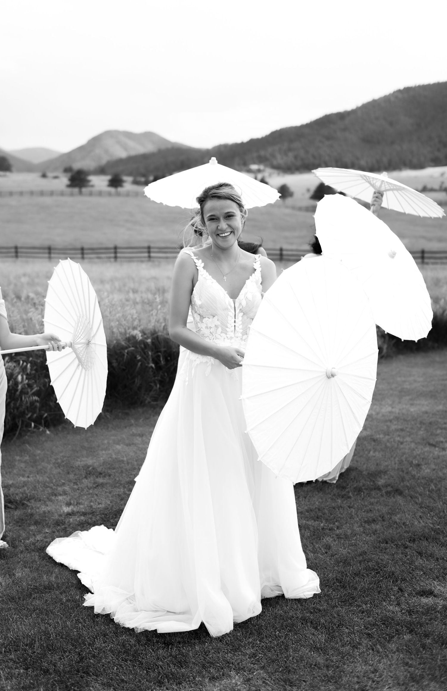 Bride in lace wedding gown smiles while holding white parasol surrounded by guests outdoors.