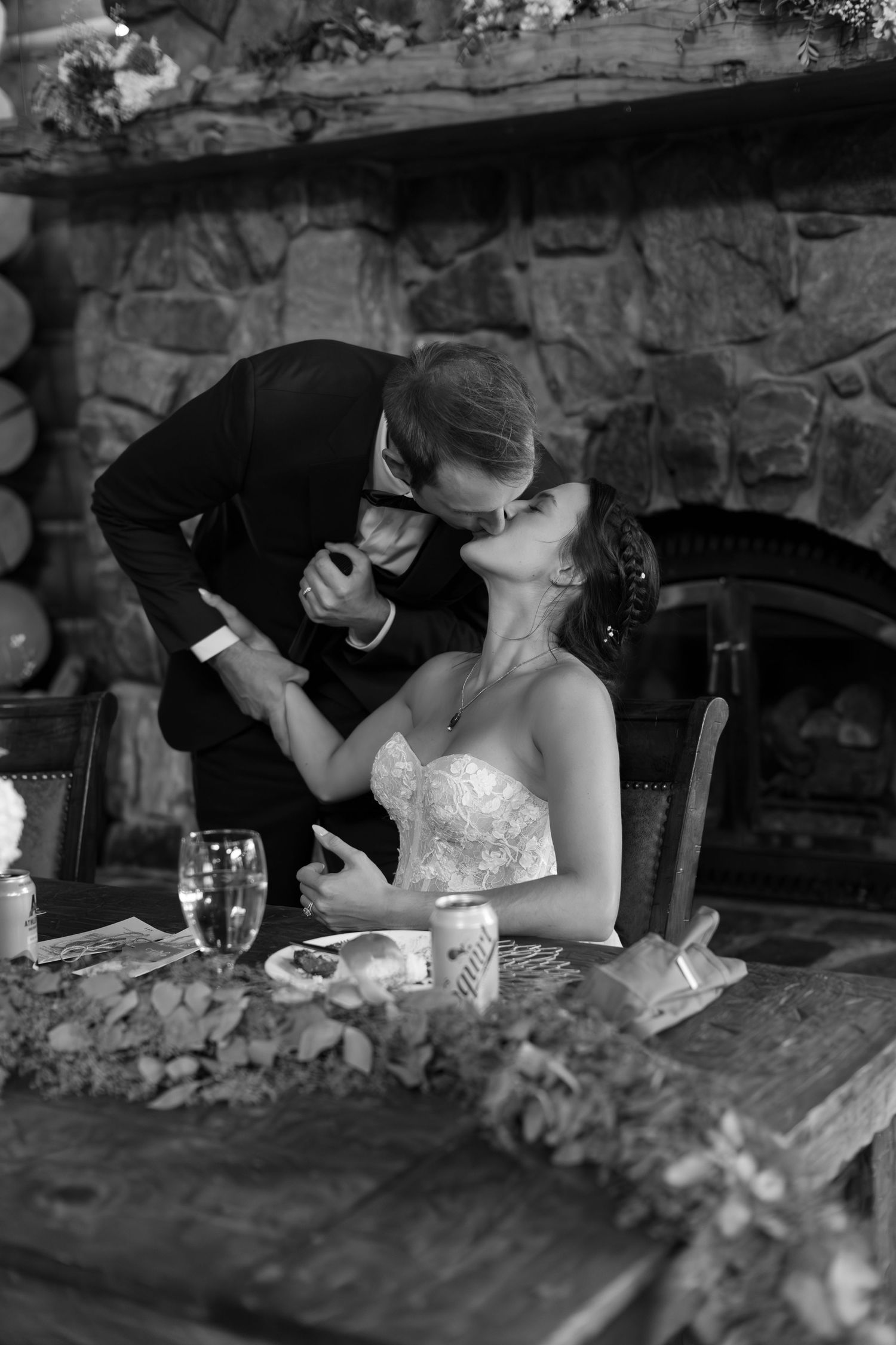Bride and groom share a romantic kiss at the sweetheart table in front of a stone fireplace.