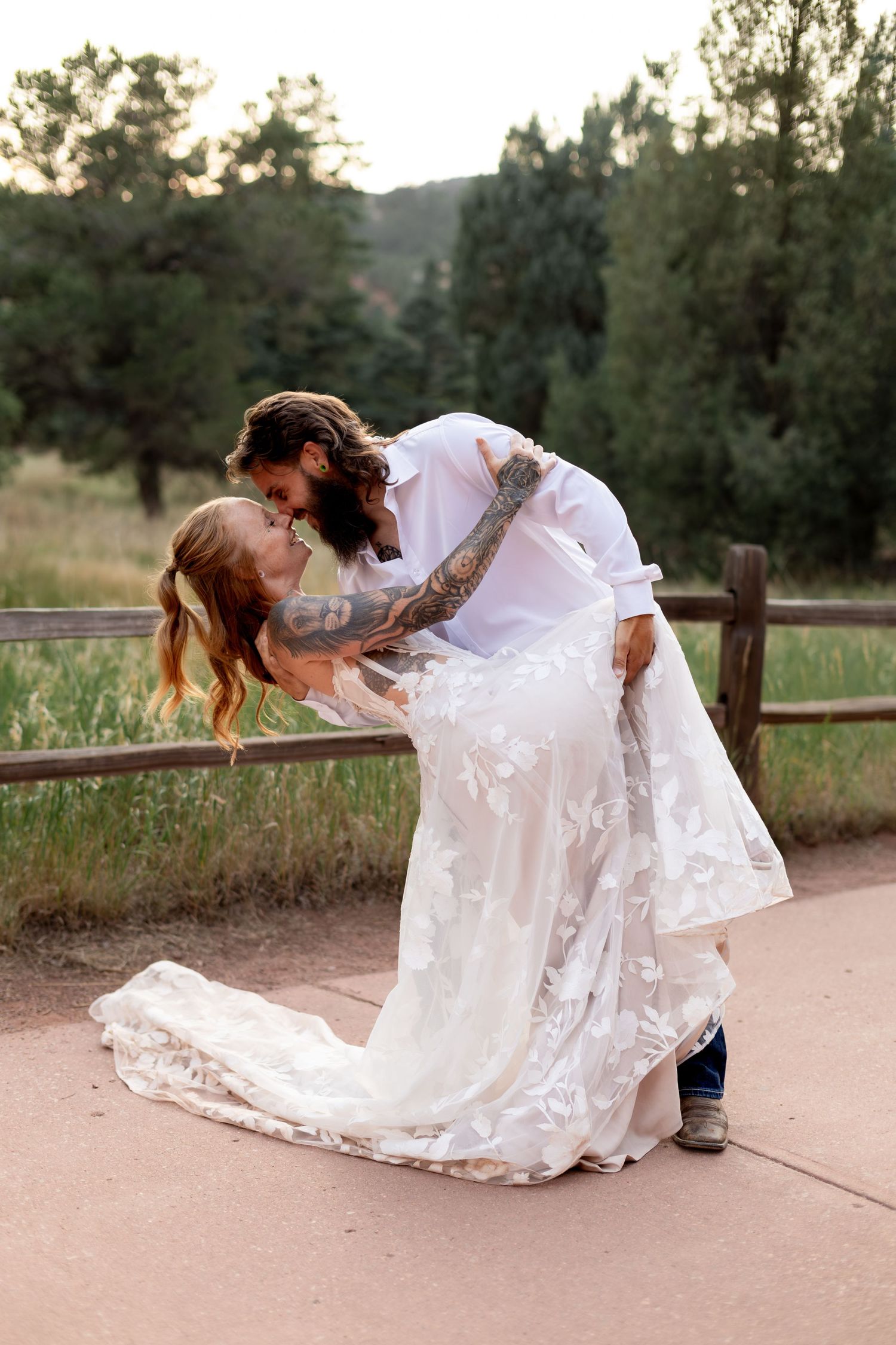 Groom with tattoos dips bride in lace wedding dress near wooden fence at a Colorado outdoor wedding venue.