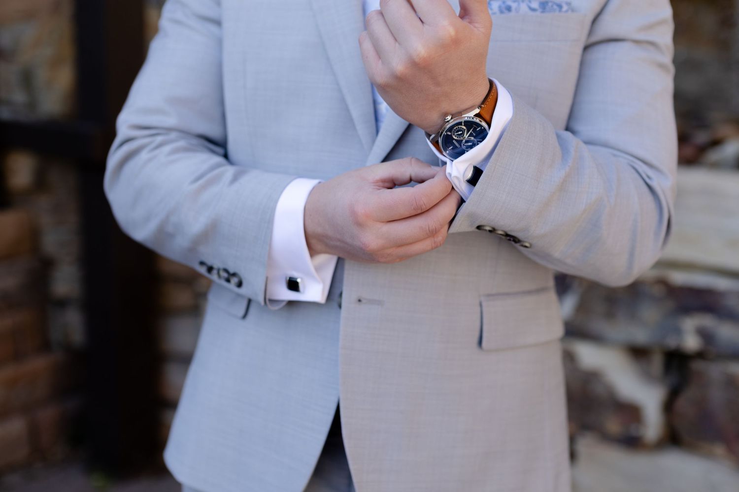Groom in light gray suit adjusting cufflinks on white dress shirt with luxury watch on wrist.