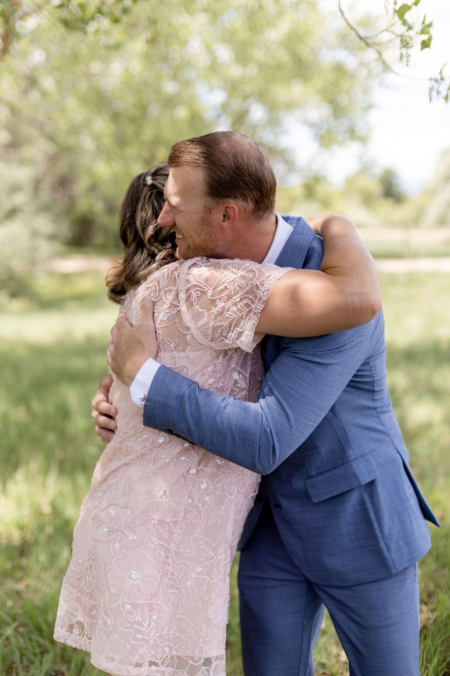Groom in blue suit embracing bride in pink lace dress outdoors at wedding ceremony.