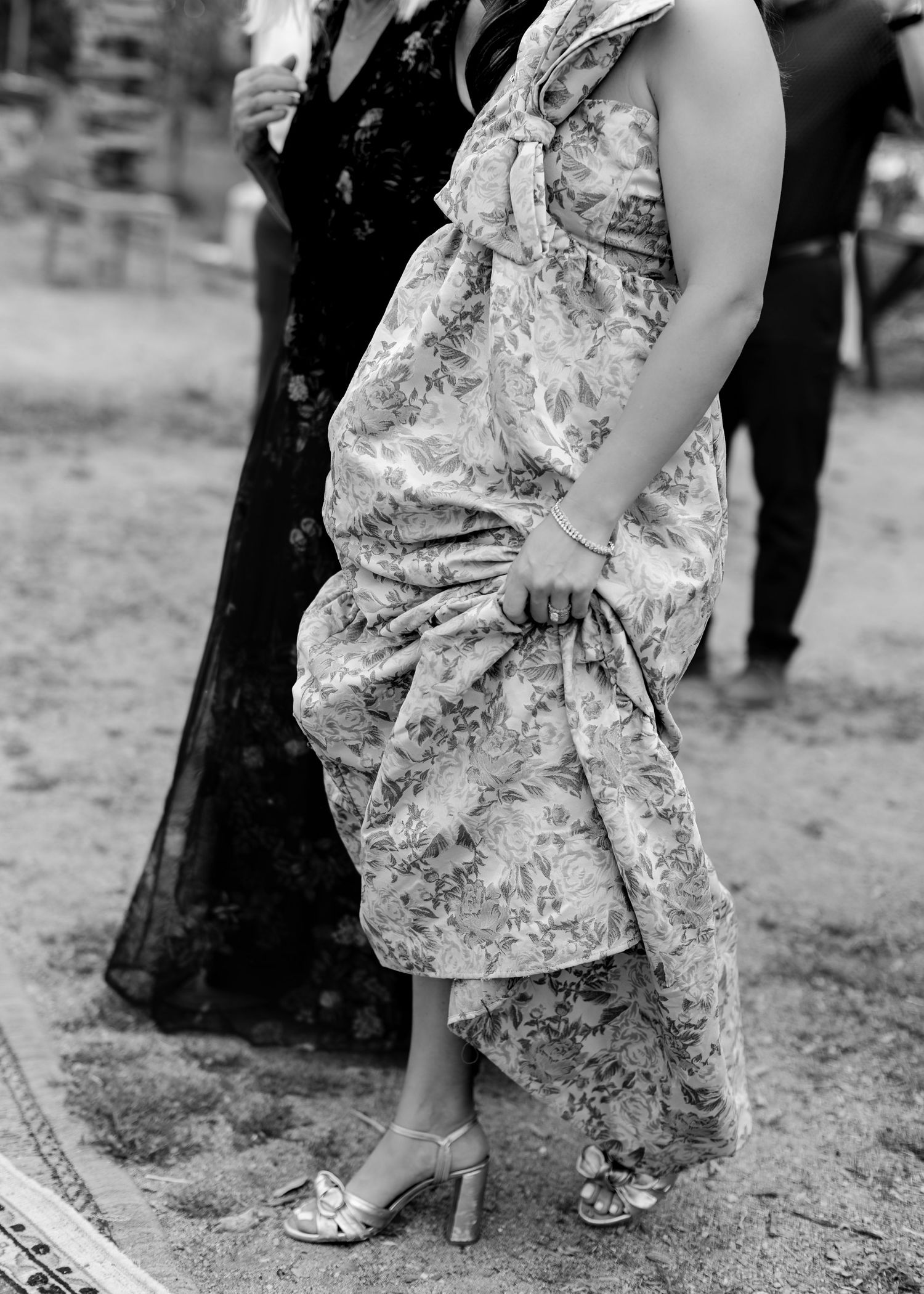 Black and white photo of pregnant woman in floral dress and metallic heels at outdoor wedding.