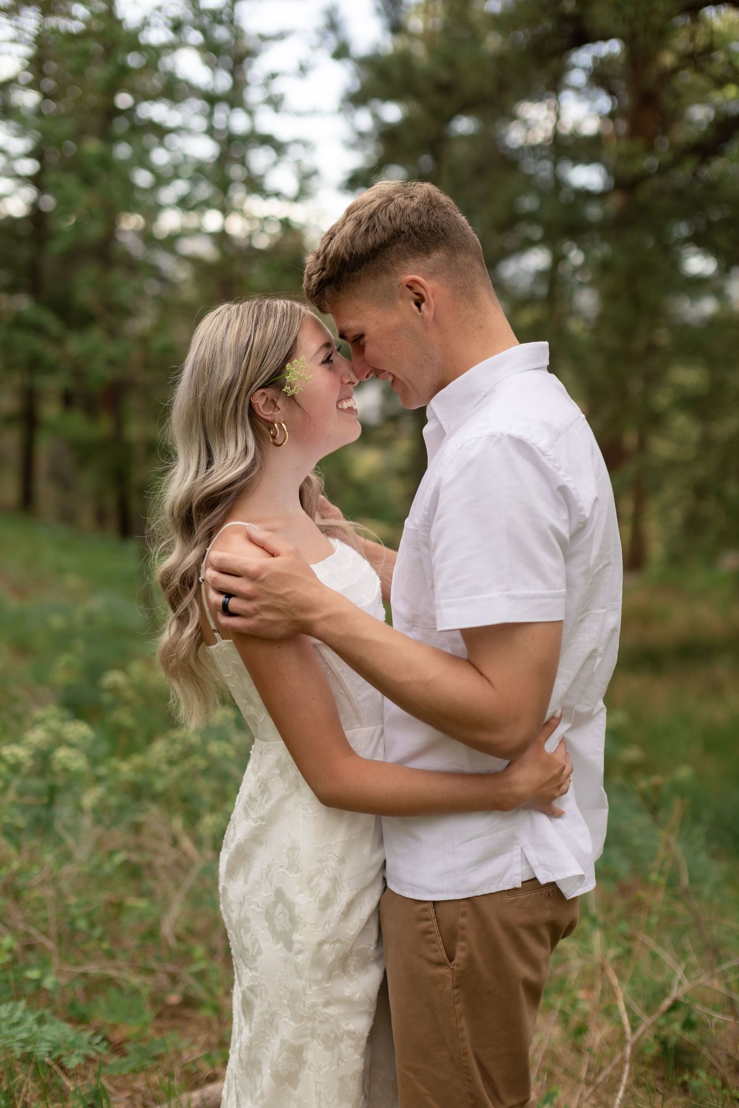 Couple embracing and sharing a romantic moment in a lush forest setting, wearing white and tan outfits.