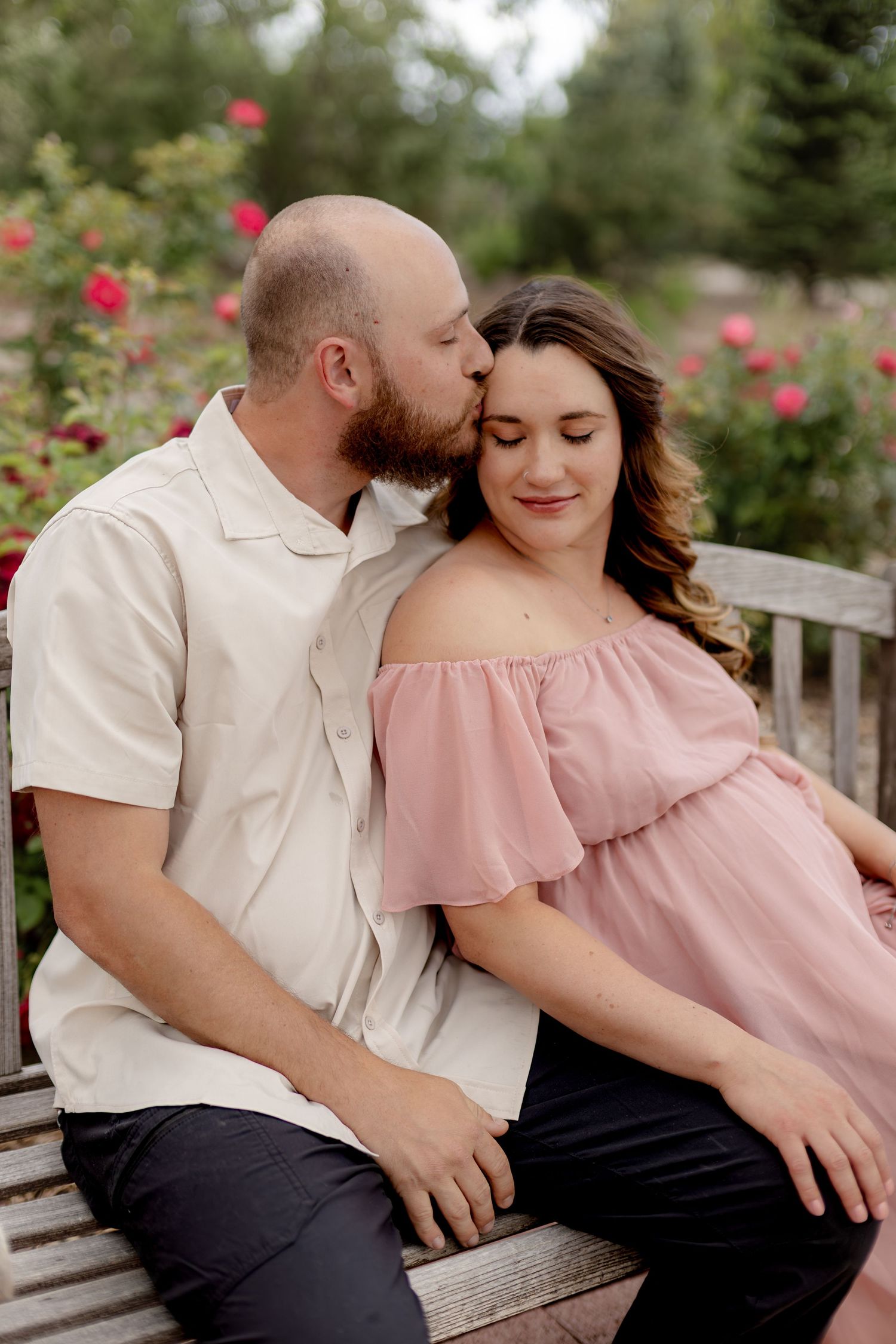 A couple sits on a bench in a rose garden, man kissing pregnant woman's temple as she smiles peacefully.