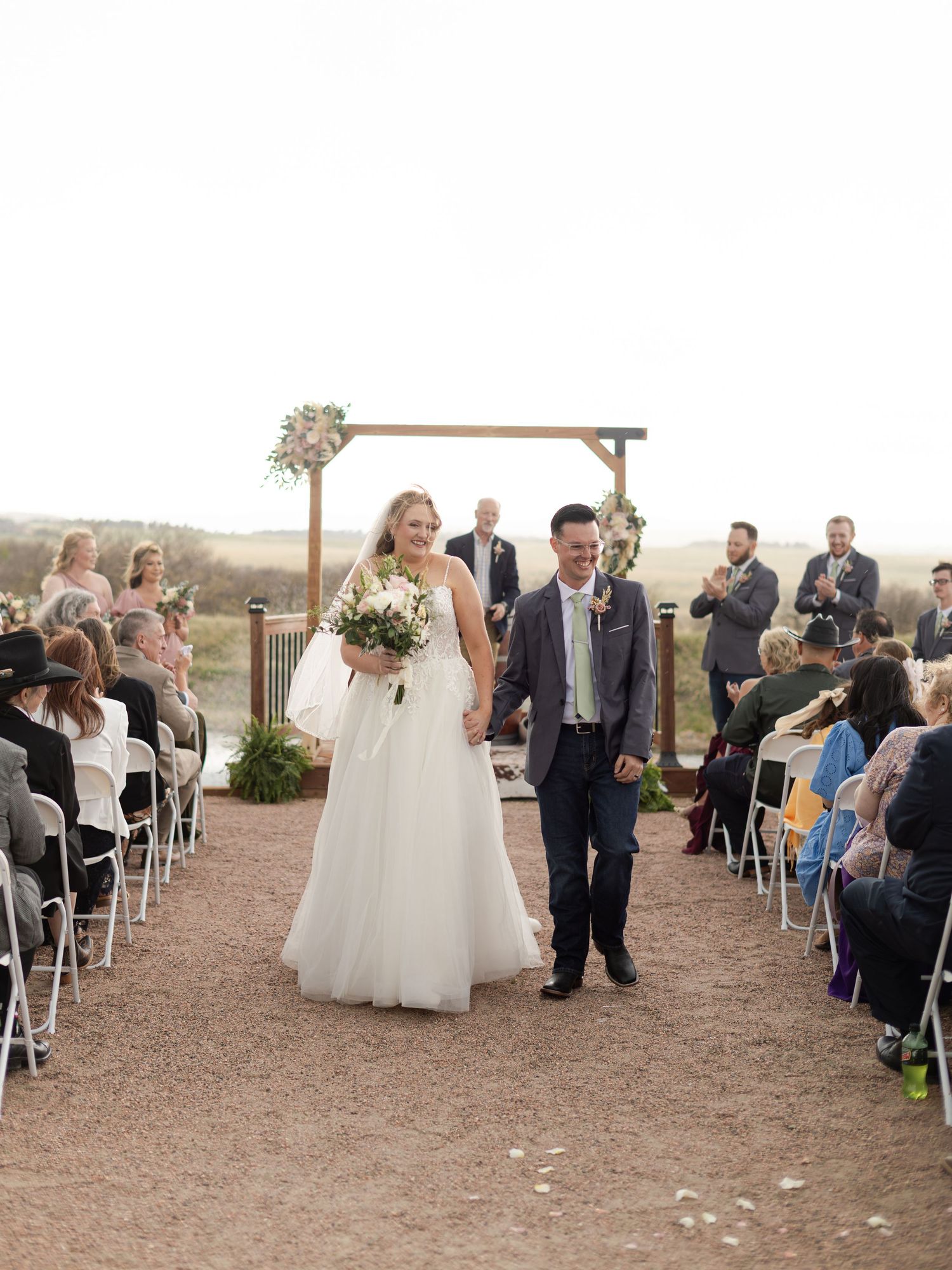 Bride and groom walk down outdoor wedding aisle smiling, guests seated on white chairs, wooden arch backdrop.