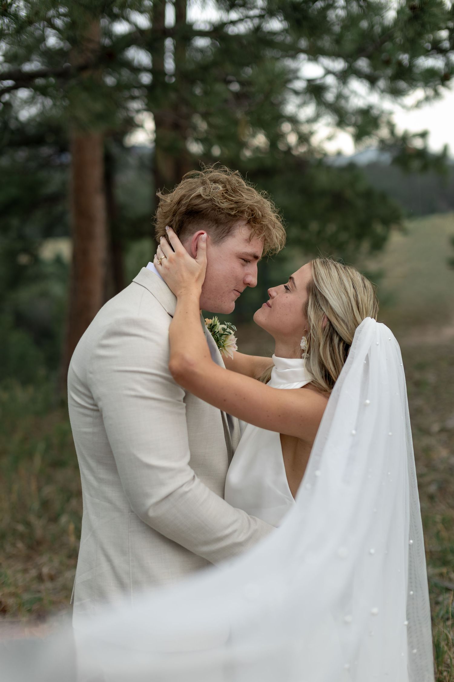 Bride and groom share tender almost-kiss moment outdoors, framed by pine trees and flowing veil.
