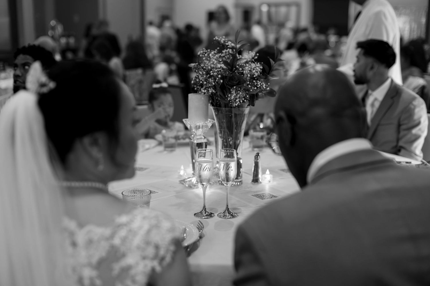 Black and white wedding reception photo showing bride, guests, and elegant floral centerpiece on decorated table.