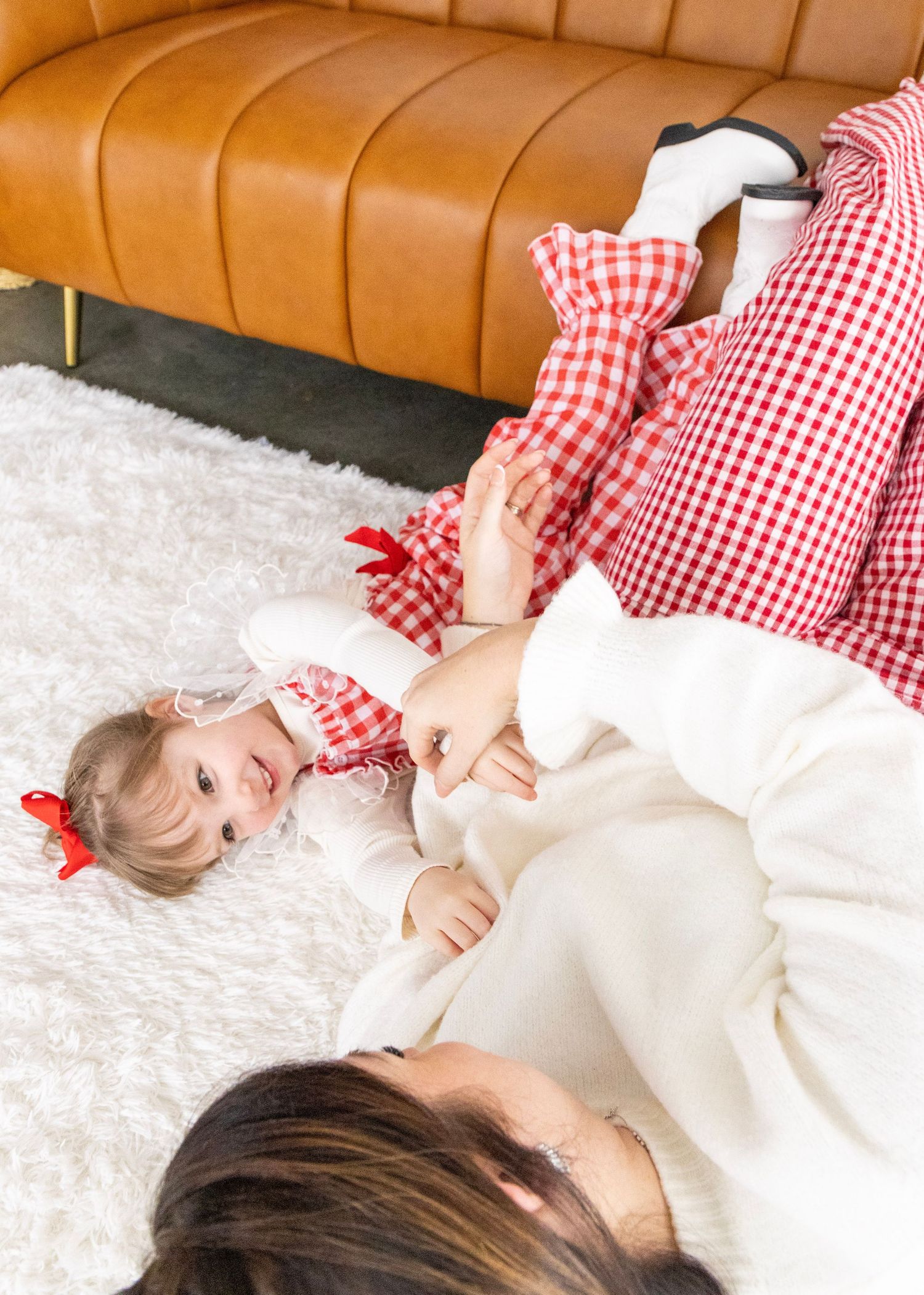 A person in red and white gingham dress lies on a white rug against a brown leather couch.