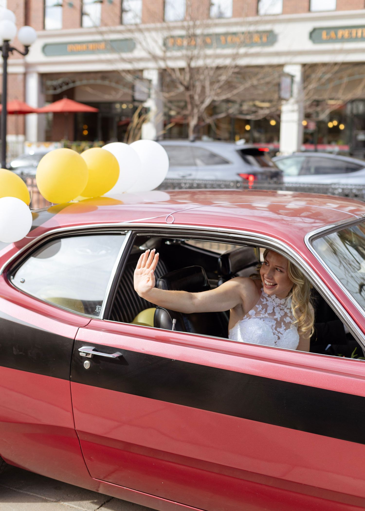 A smiling bride in a white dress waves from a red classic car decorated with gold and white balloons.