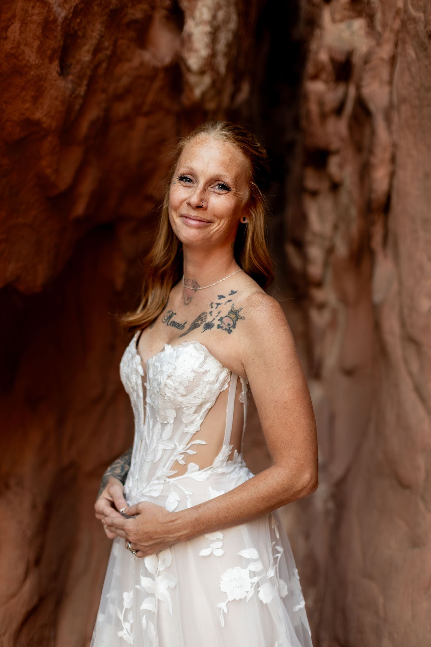 A tattooed bride in a white floral lace gown poses gracefully in a red rock canyon slot.