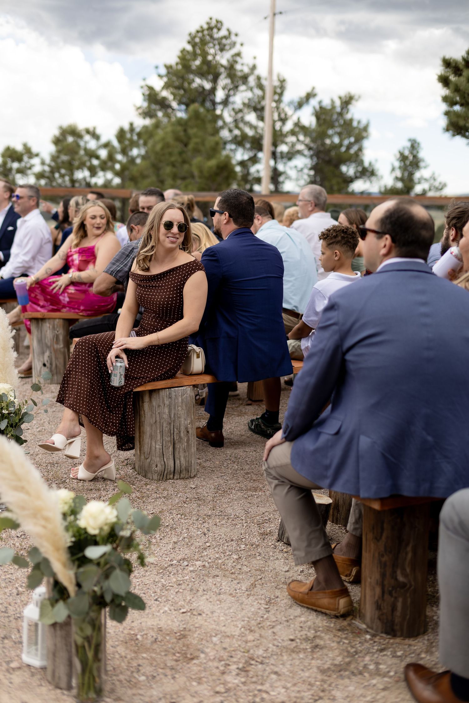 Wedding guests seated on wooden benches at outdoor rustic ceremony with white floral aisle decorations.
