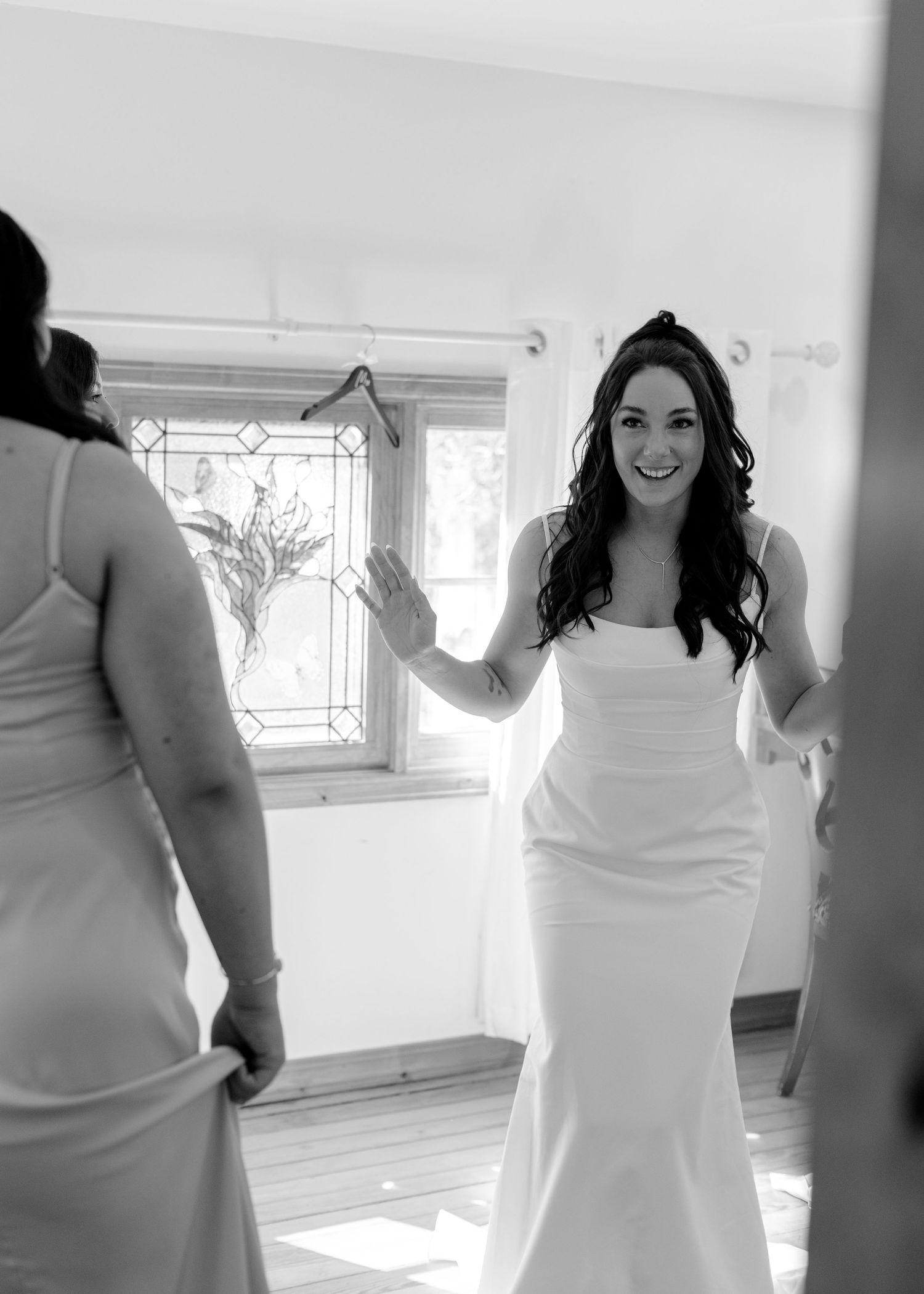 Black and white photo of a smiling bride in a sleek white gown admiring herself in a mirror before her wedding.
