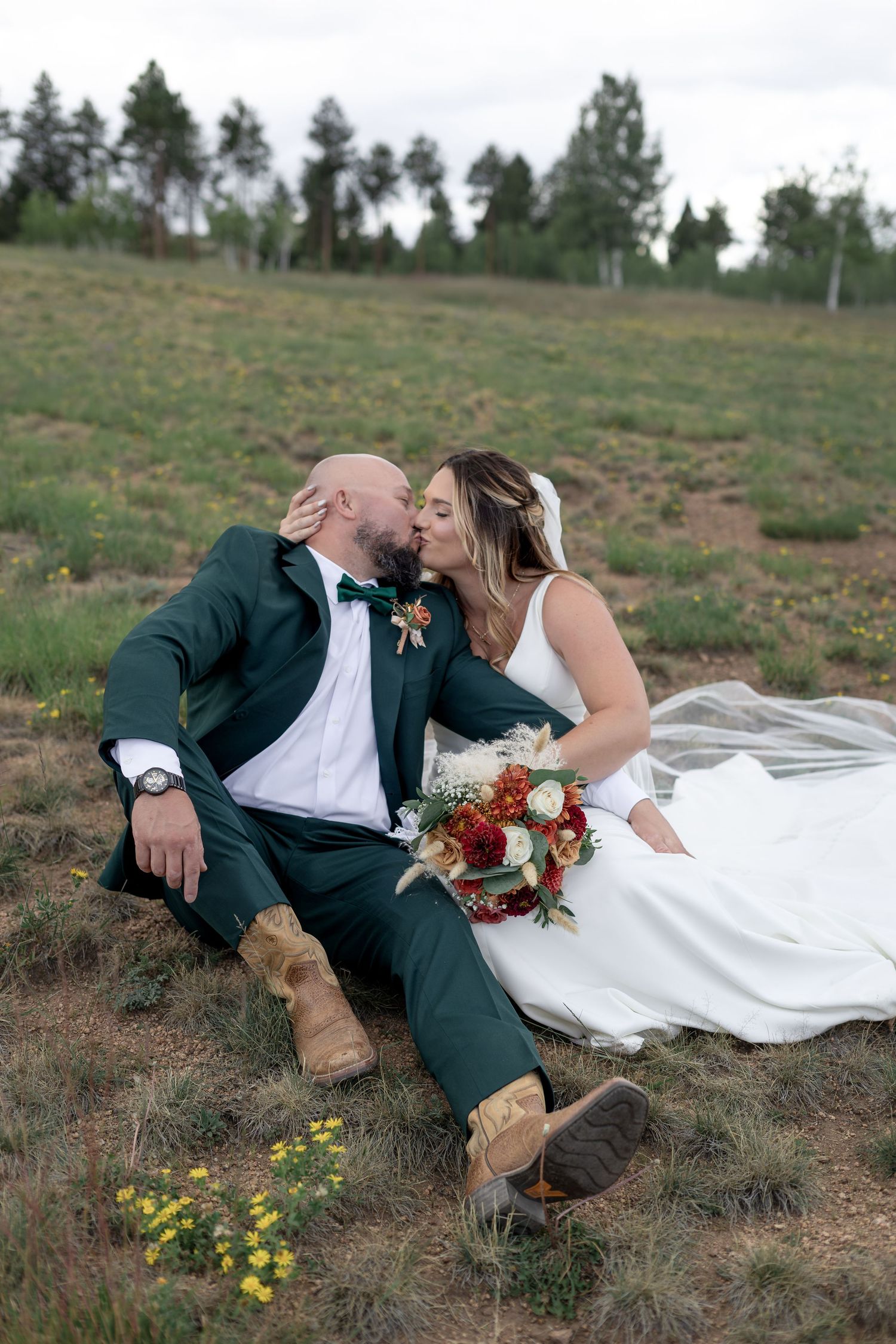 Bride and groom sitting on a grassy hillside kissing, wearing white dress and green suit with cowboy boots, holding red bouquet.