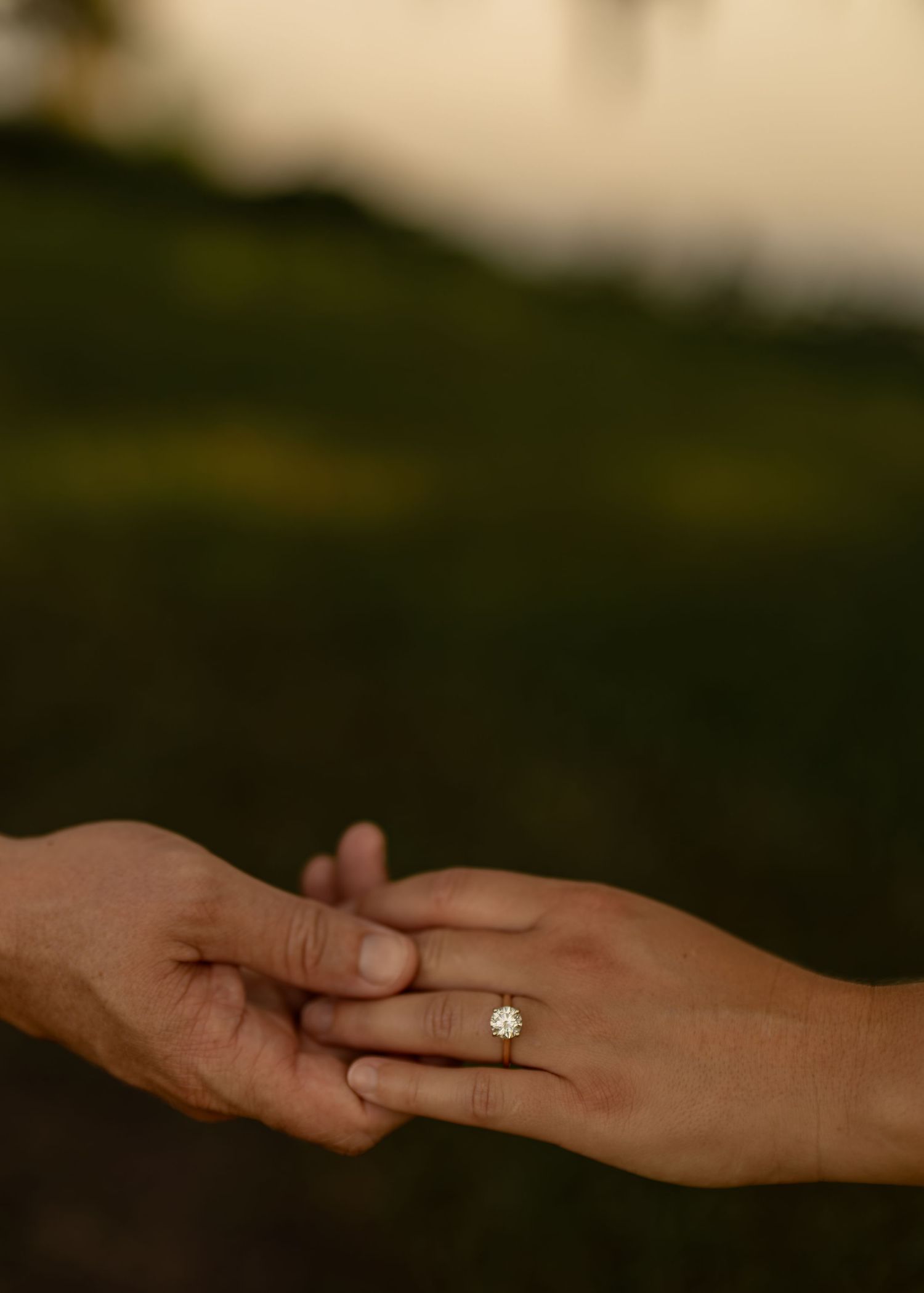 Two hands gently touching, one adorned with a gold solitaire diamond engagement ring, set against a blurred green outdoor background.