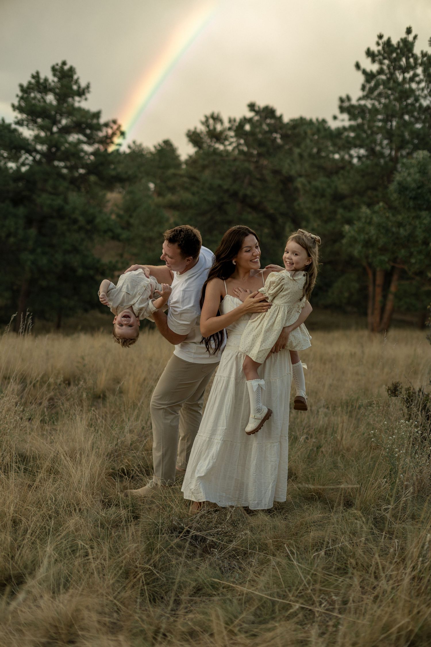 Happy family with two children playing in a field with a rainbow and pine trees in the background.