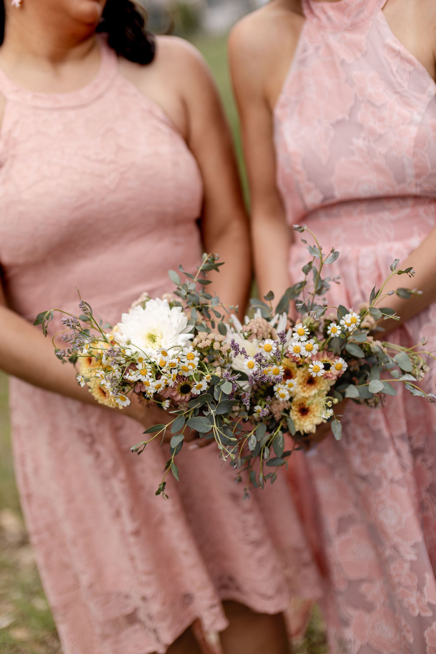 Two bridesmaids in pink lace dresses holding wildflower bouquets with daisies, eucalyptus, and yellow blooms at a wedding.