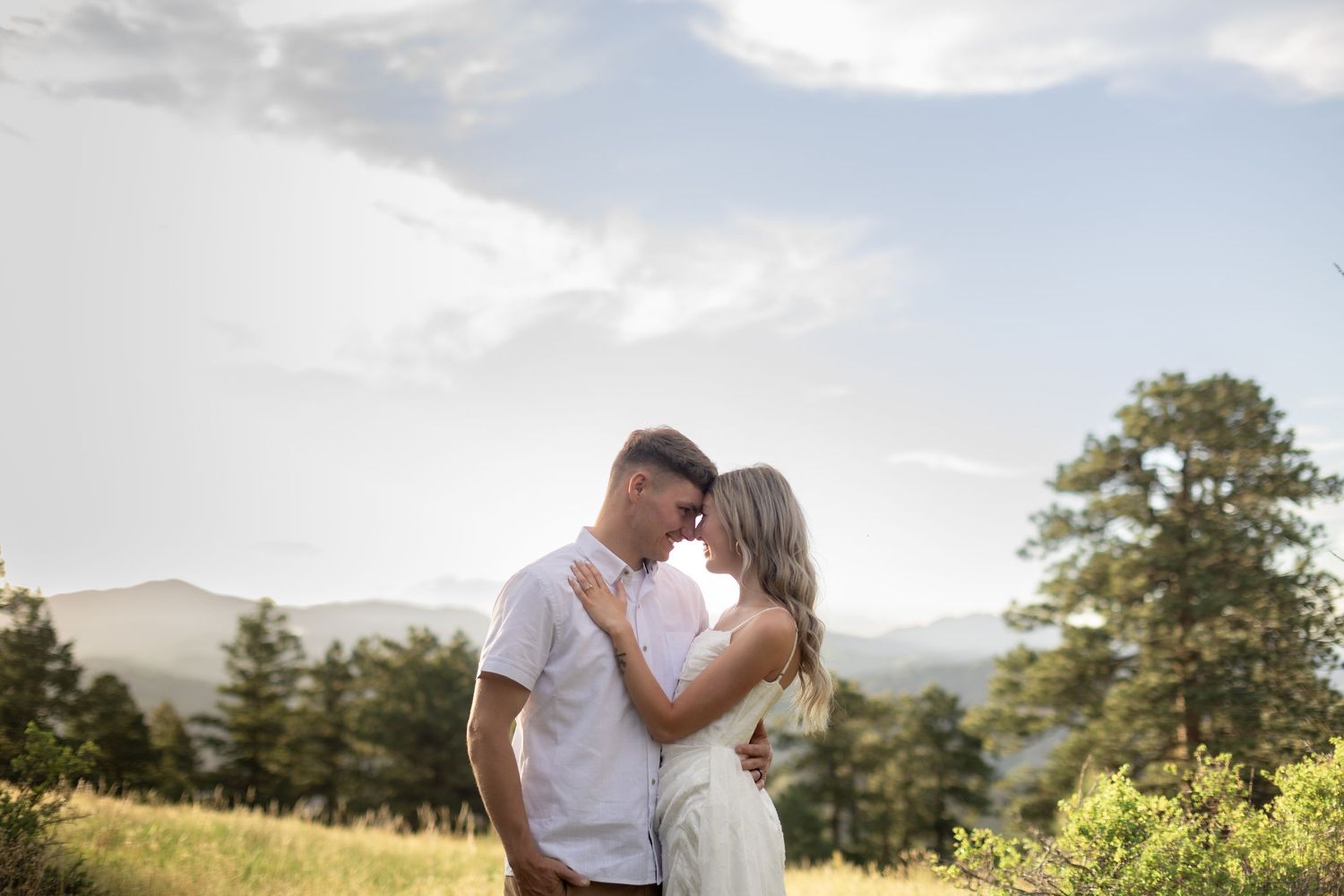 Couple embracing on a mountain meadow with pine trees and cloudy sky in a romantic outdoor engagement photo.