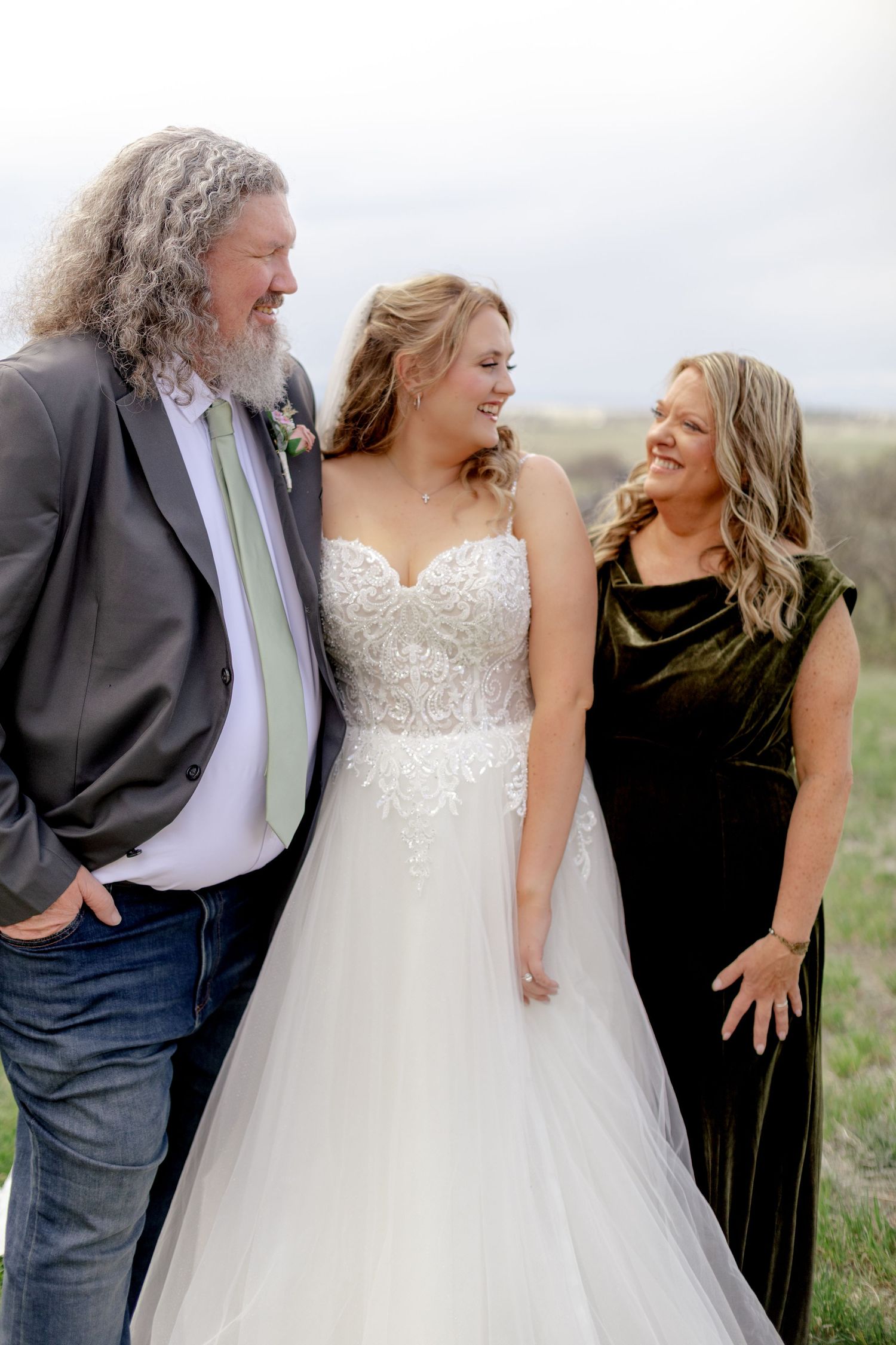 Bride in white lace gown smiling with parents outdoors on her wedding day.