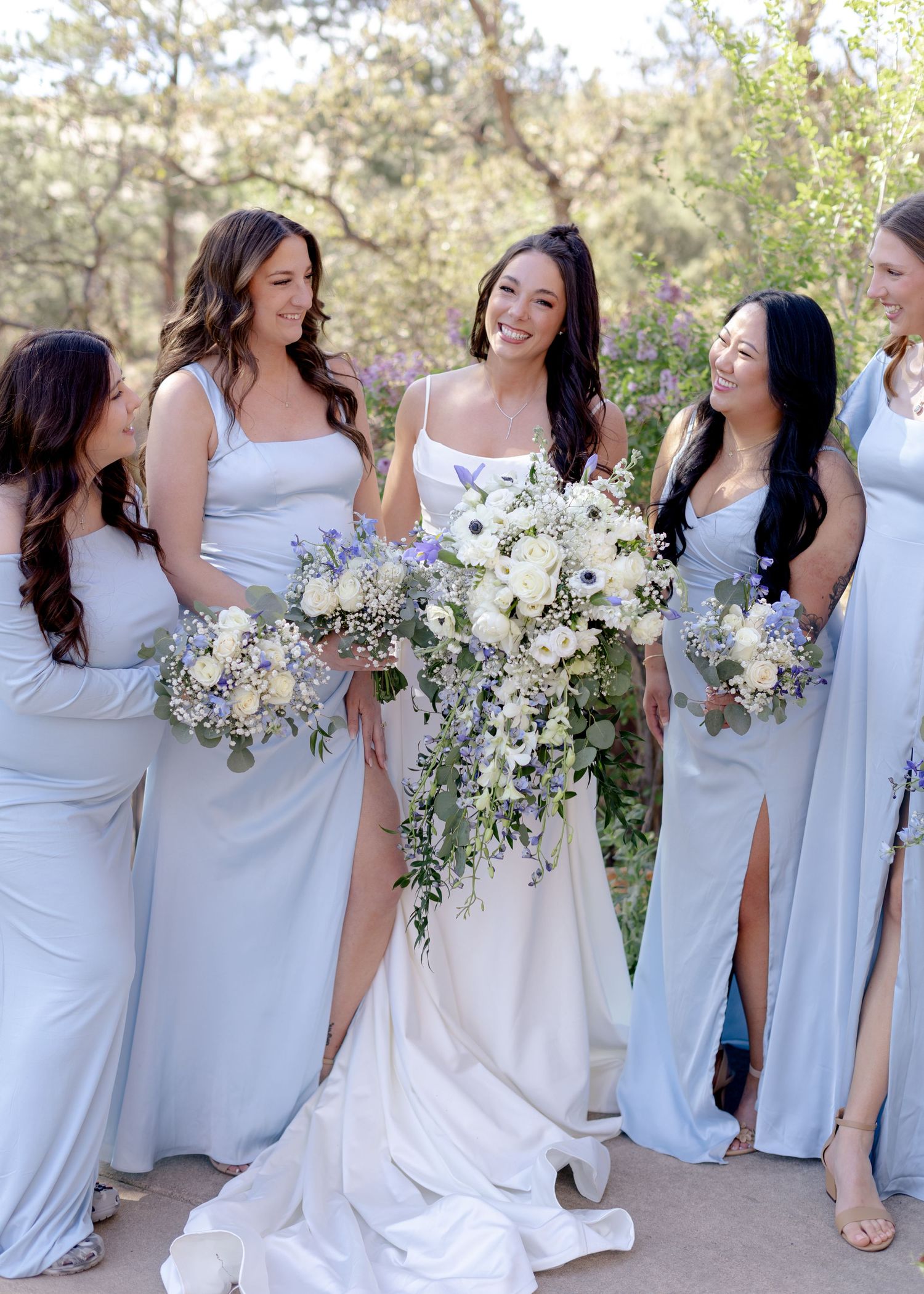 Bride in white gown smiles with bridesmaids in light blue dresses holding white and blue floral bouquets outdoors.