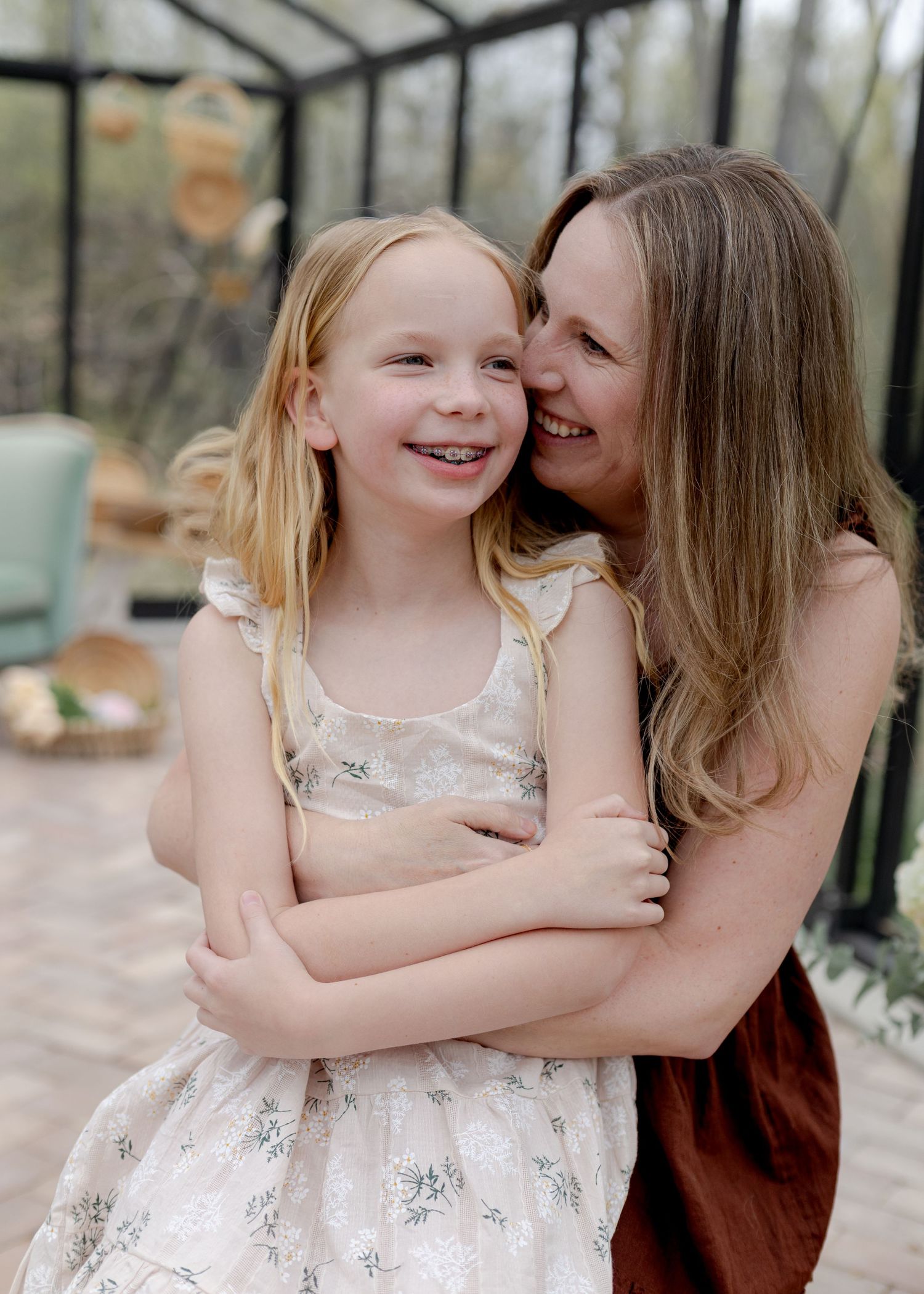A woman hugging a young blonde girl in a floral dress, both smiling warmly in a bright greenhouse setting.