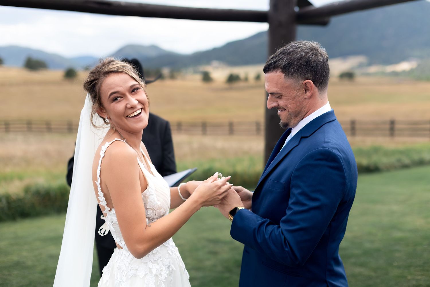 Bride laughing joyfully while exchanging rings with groom in blue suit at outdoor wedding ceremony with mountain backdrop.
