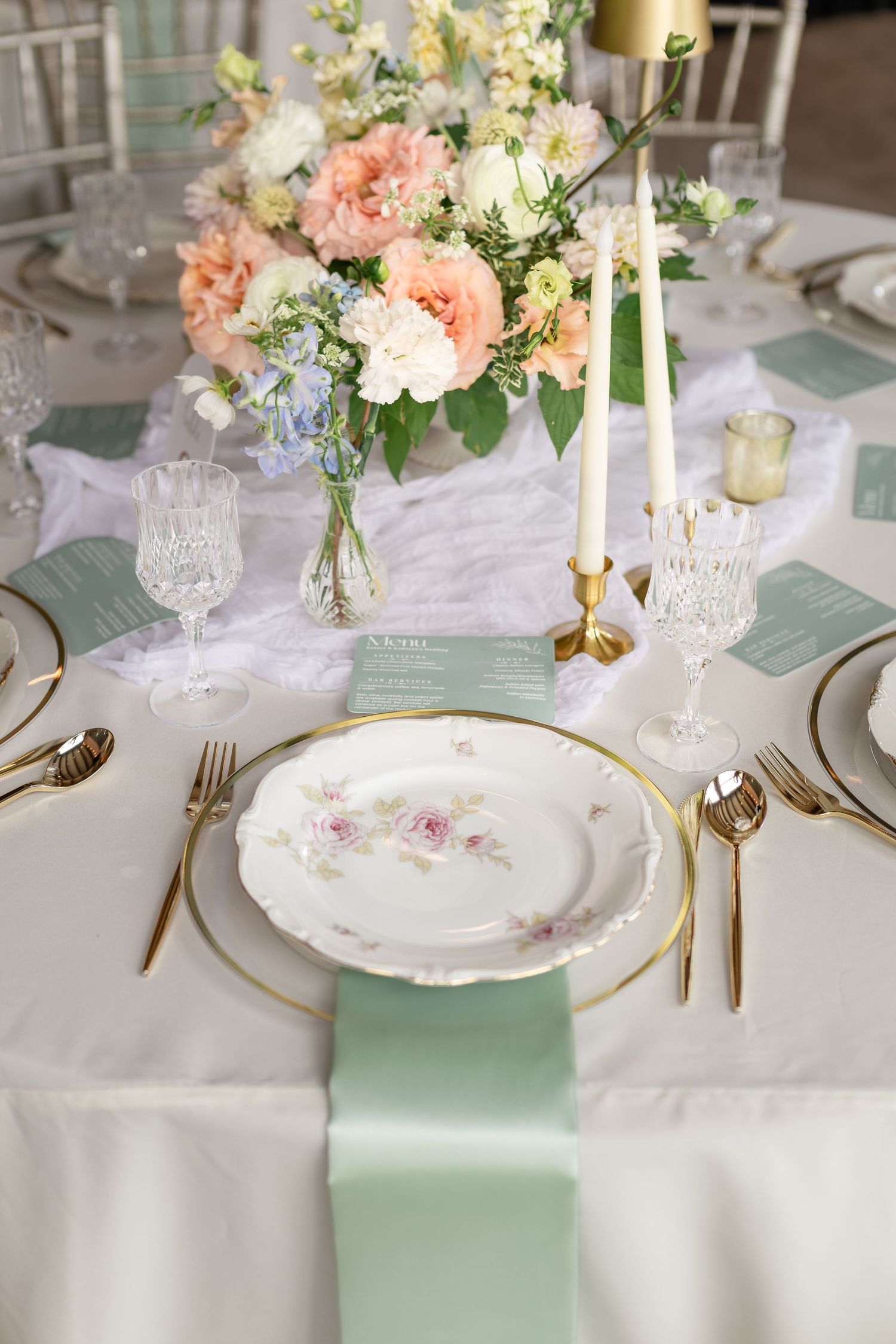 Elegant wedding table setting with mint green napkins, gold cutlery, floral china, crystal glasses, and a peach floral centerpiece.