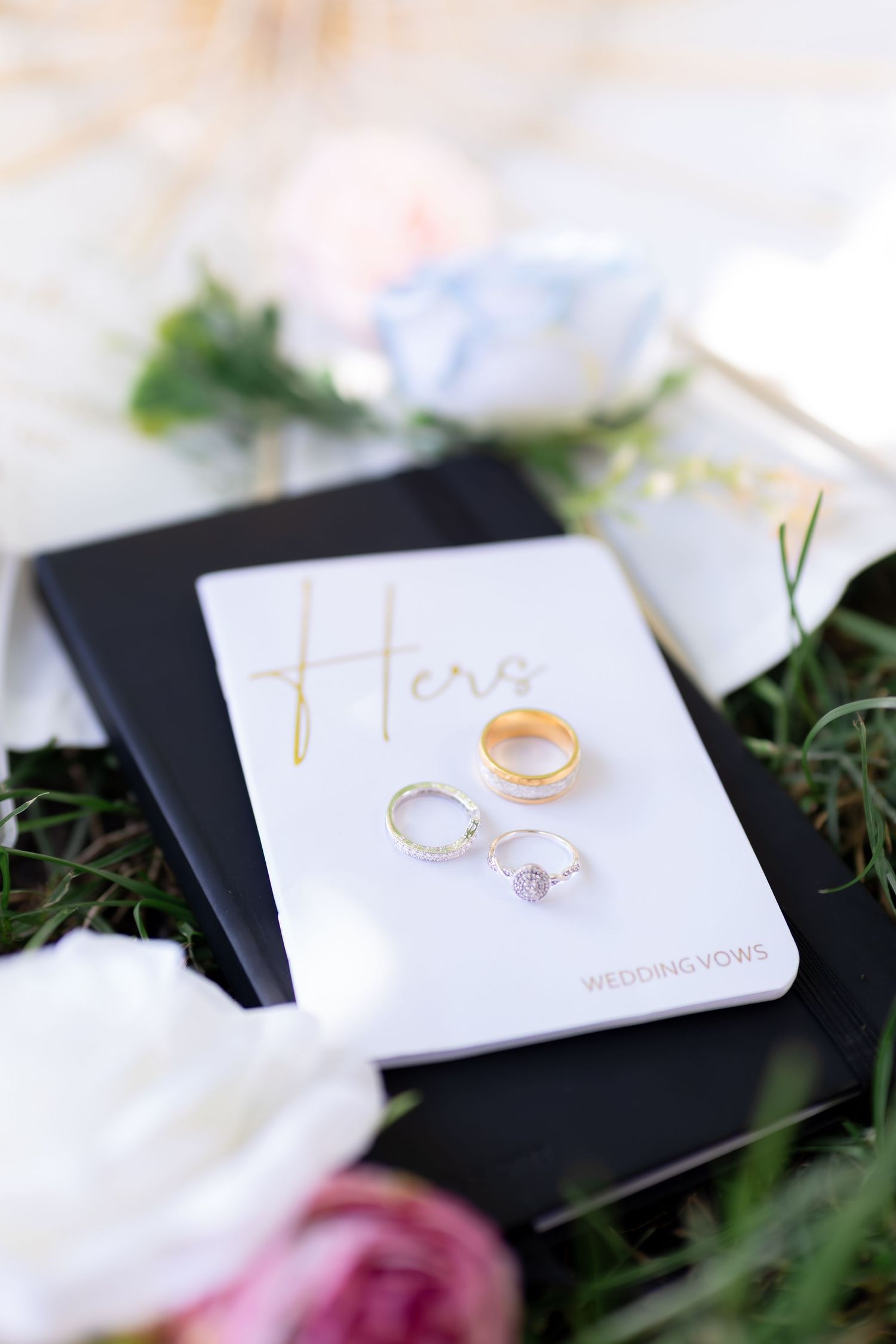 Wedding rings and engagement ring resting on a white vows book labeled Hers, surrounded by flowers.
