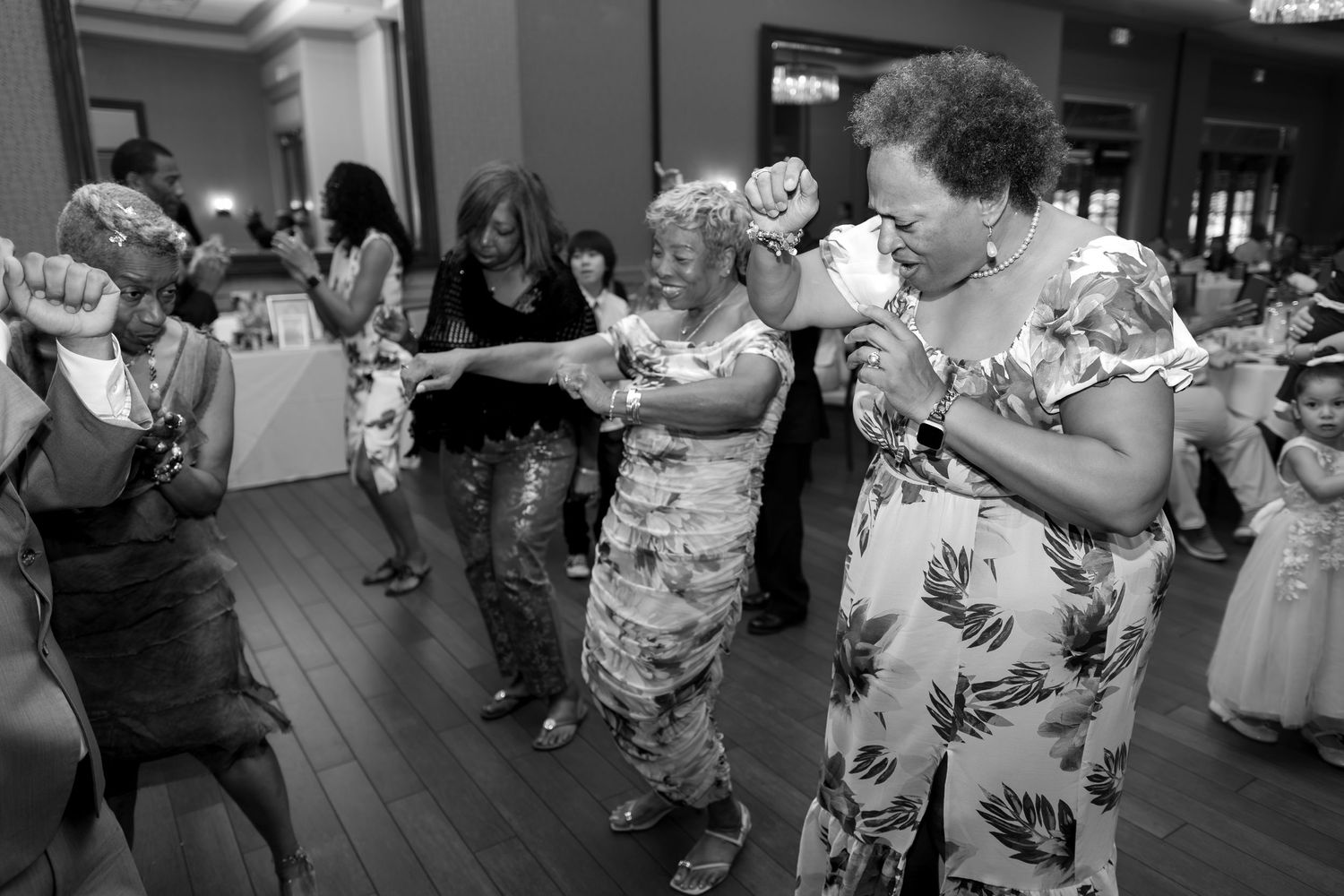 Women dancing joyfully at an indoor celebration event, wearing festive attire on a wooden dance floor.
