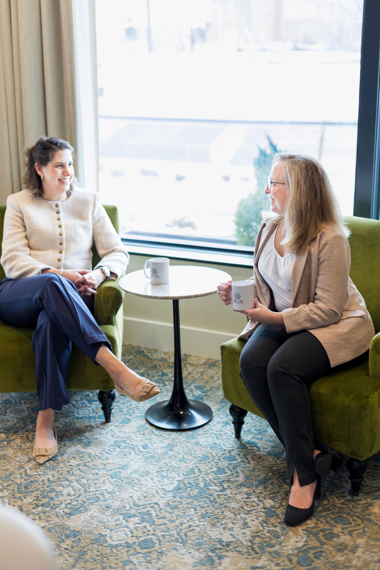 Two people sit in plush green chairs having a conversation by a window in an elegant room with patterned carpet.