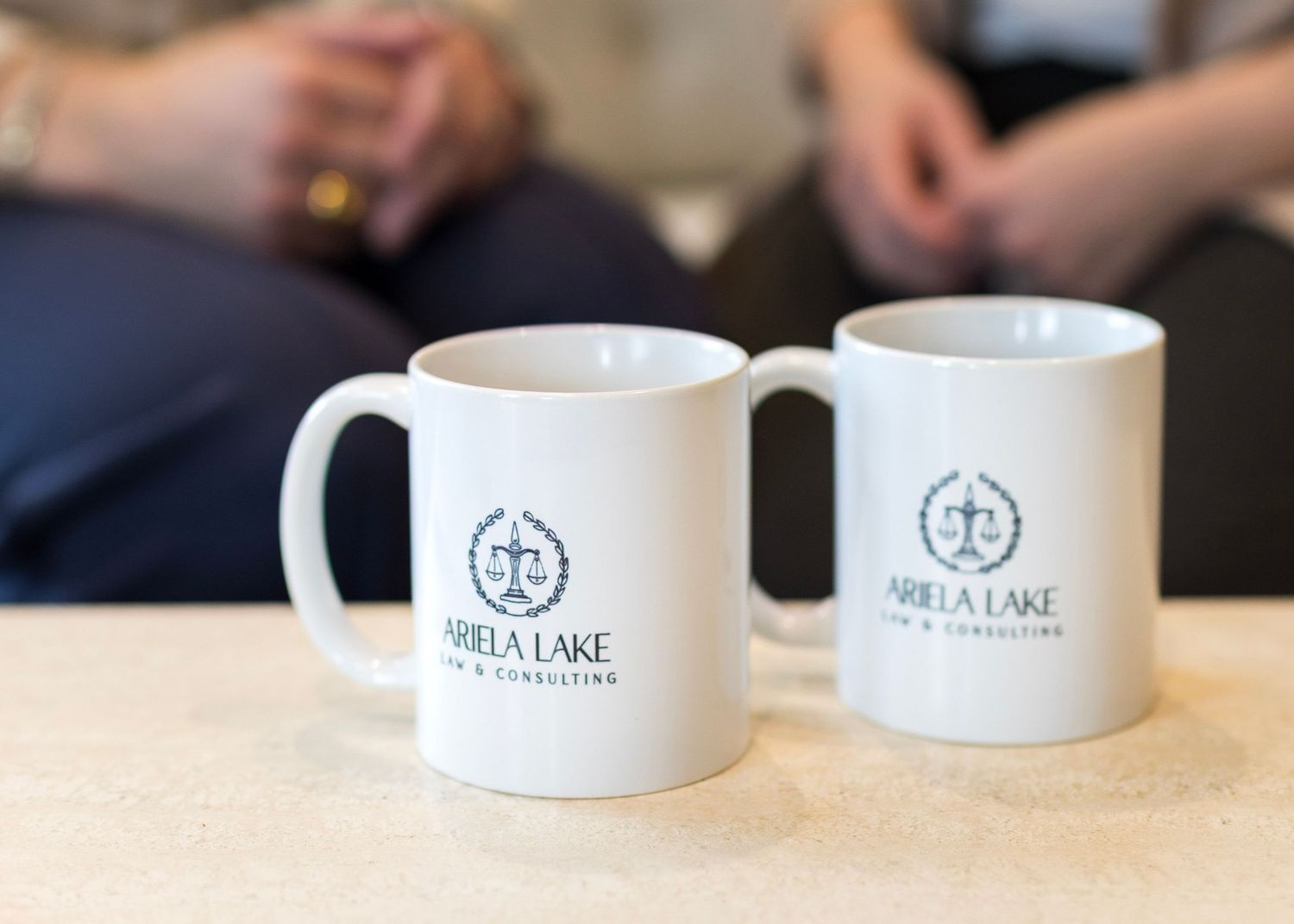 Two white coffee mugs with Della Lake logo branding sit on a wooden table during a meeting.