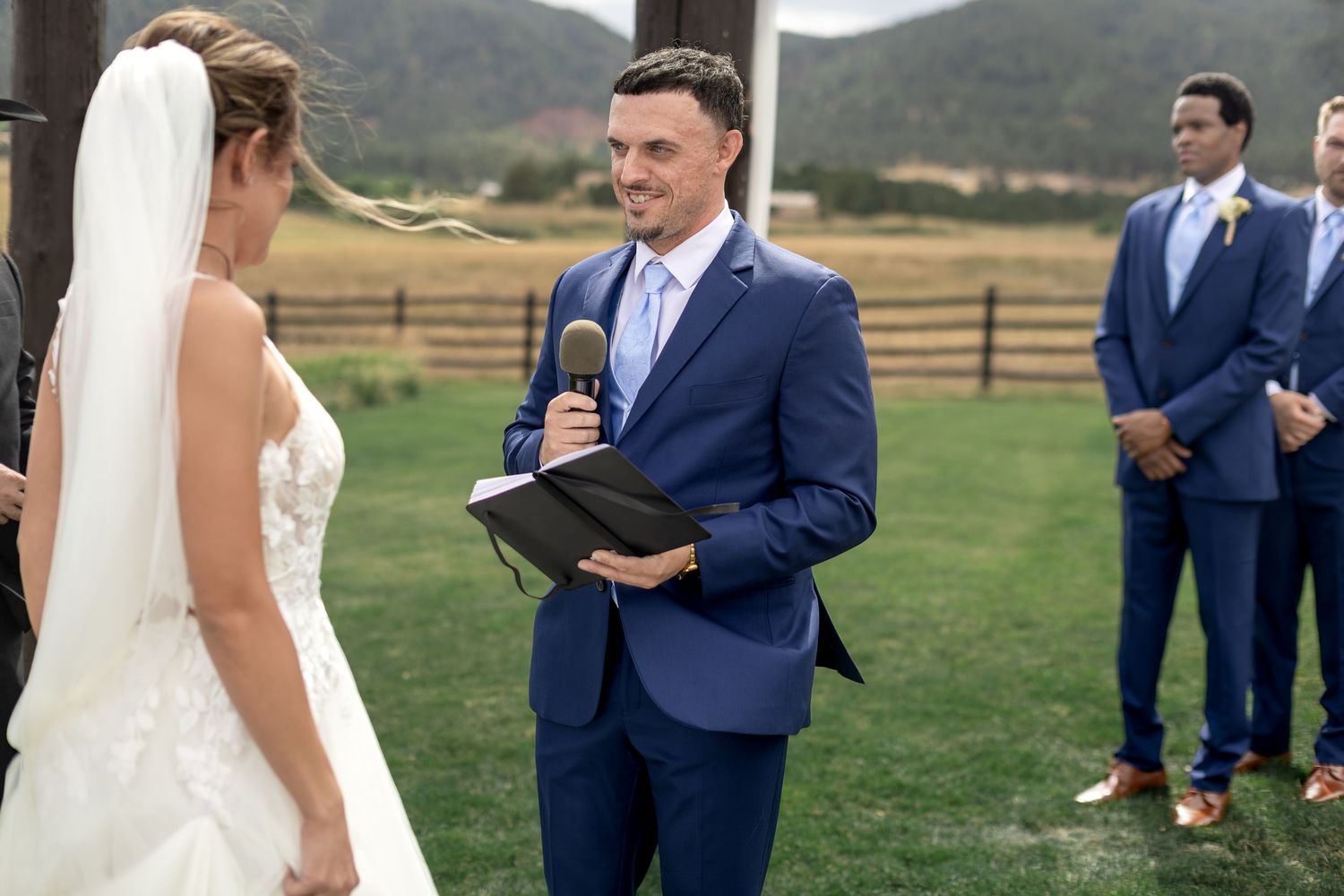 Groom in navy suit reads vows to bride in white wedding dress during outdoor ceremony with mountain backdrop.