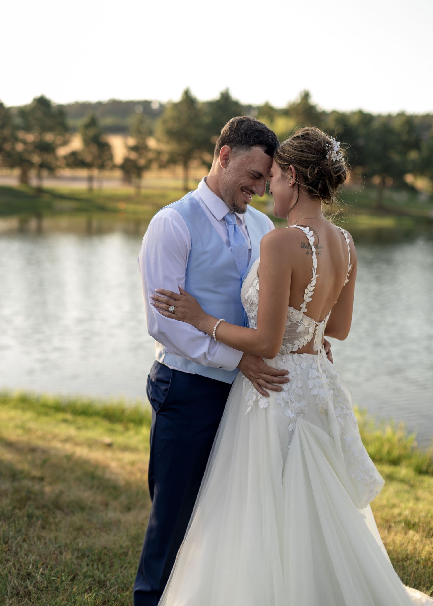 Bride and groom share an intimate moment touching foreheads, smiling by a tranquil pond at sunset on their wedding day.