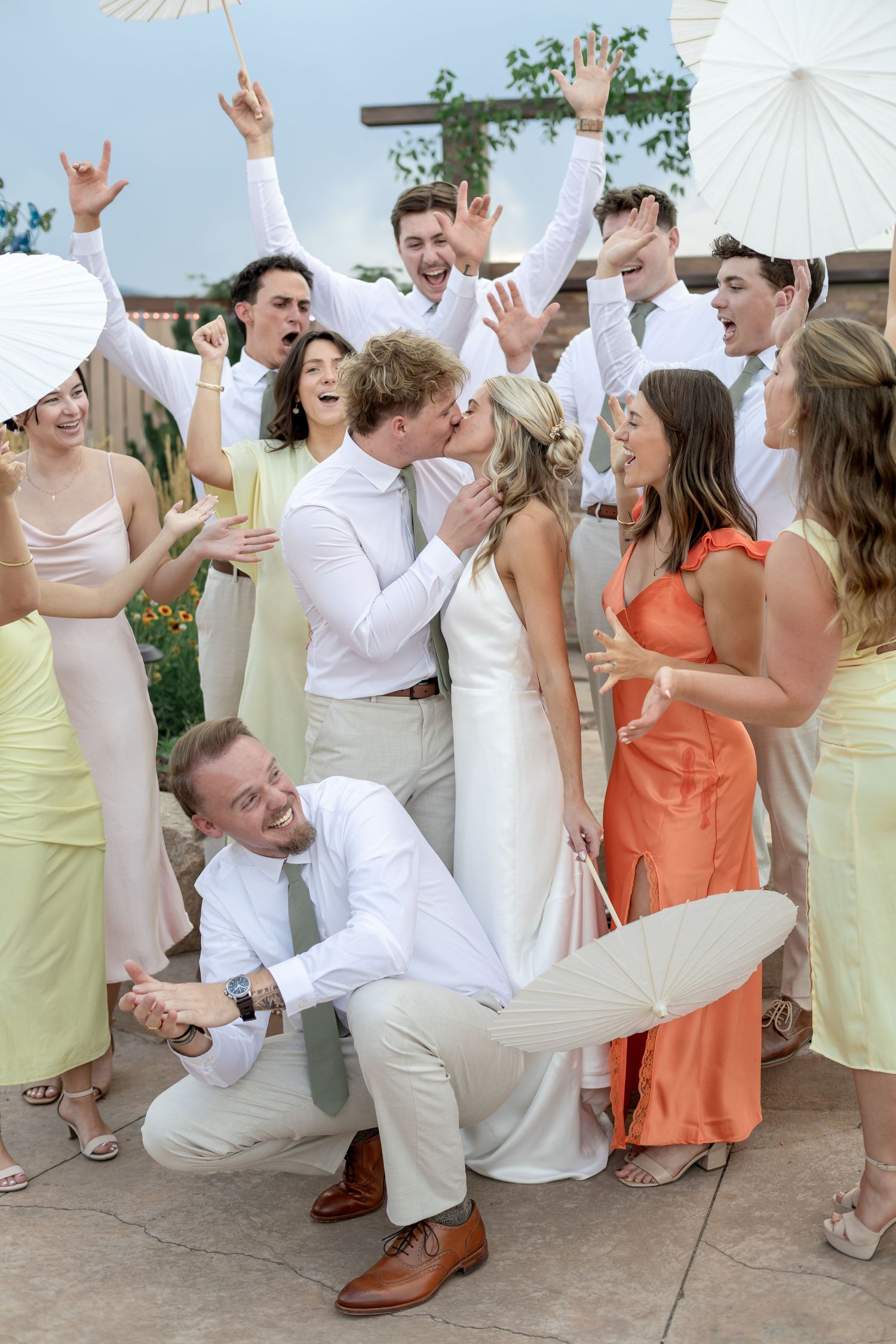 Wedding party celebrates as bride and groom kiss outdoors, surrounded by cheering guests in pastel and orange attire.