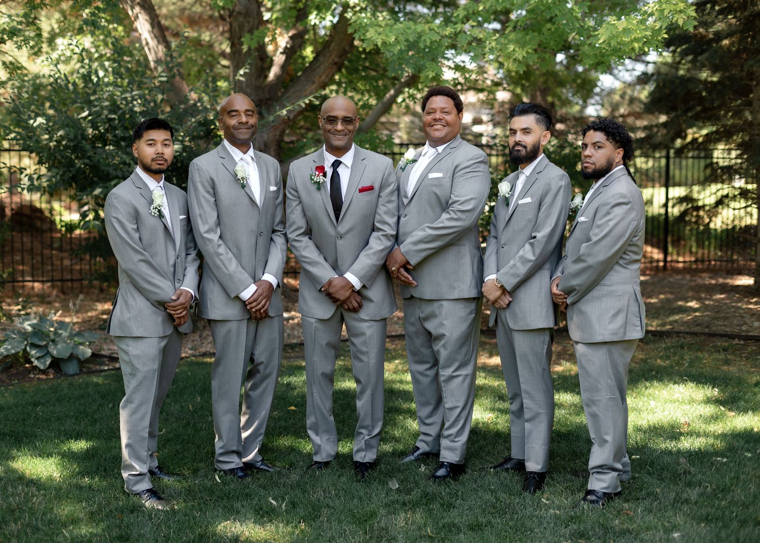 Six groomsmen in matching light gray suits pose outdoors on green grass for a wedding party photo.