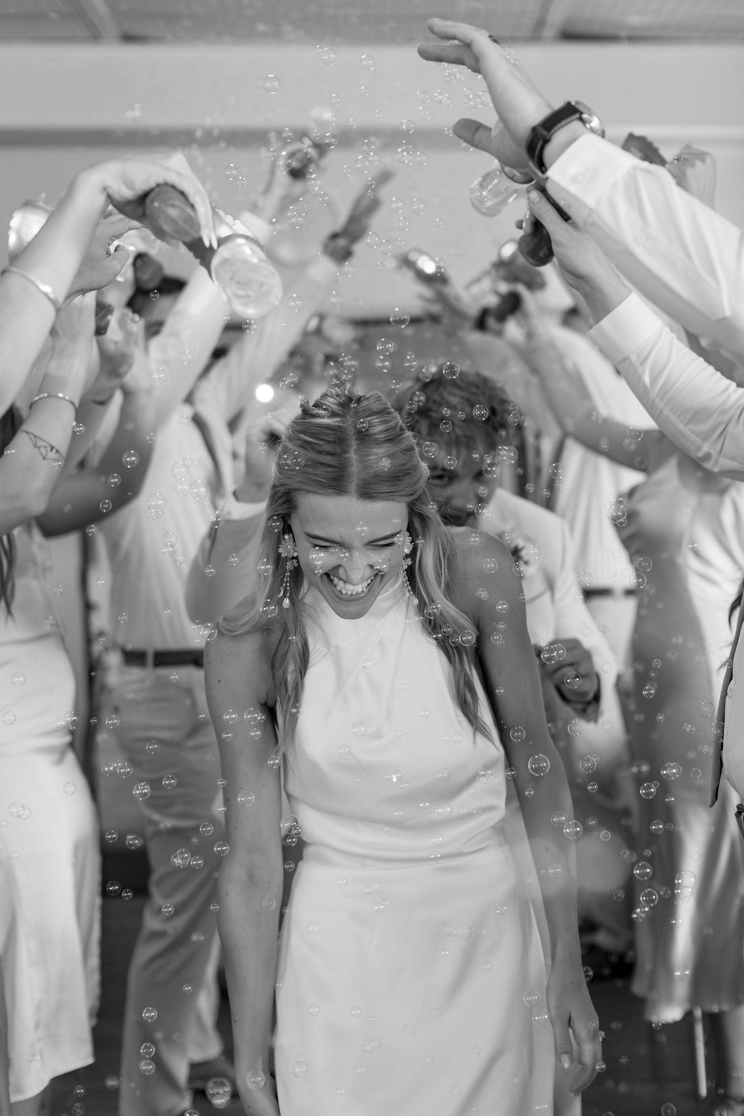 Bride laughing joyfully as guests blow bubbles during wedding reception send-off in black and white photo.