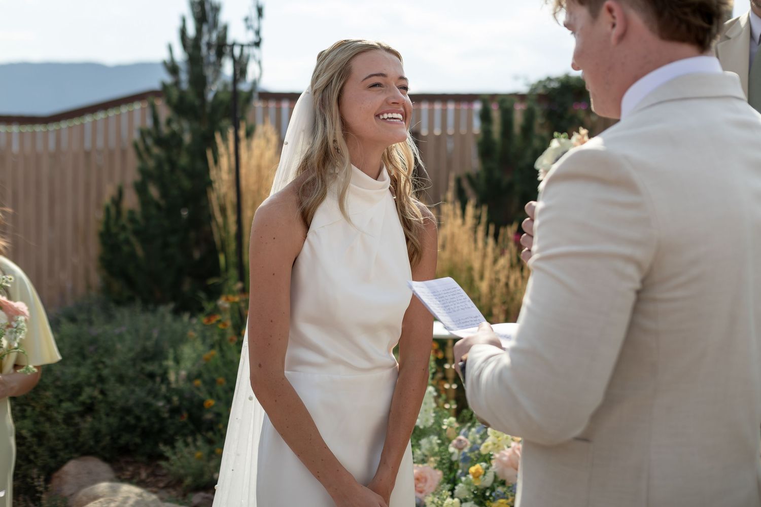 Bride in white sleeveless gown smiling at groom during outdoor wedding ceremony with garden backdrop.