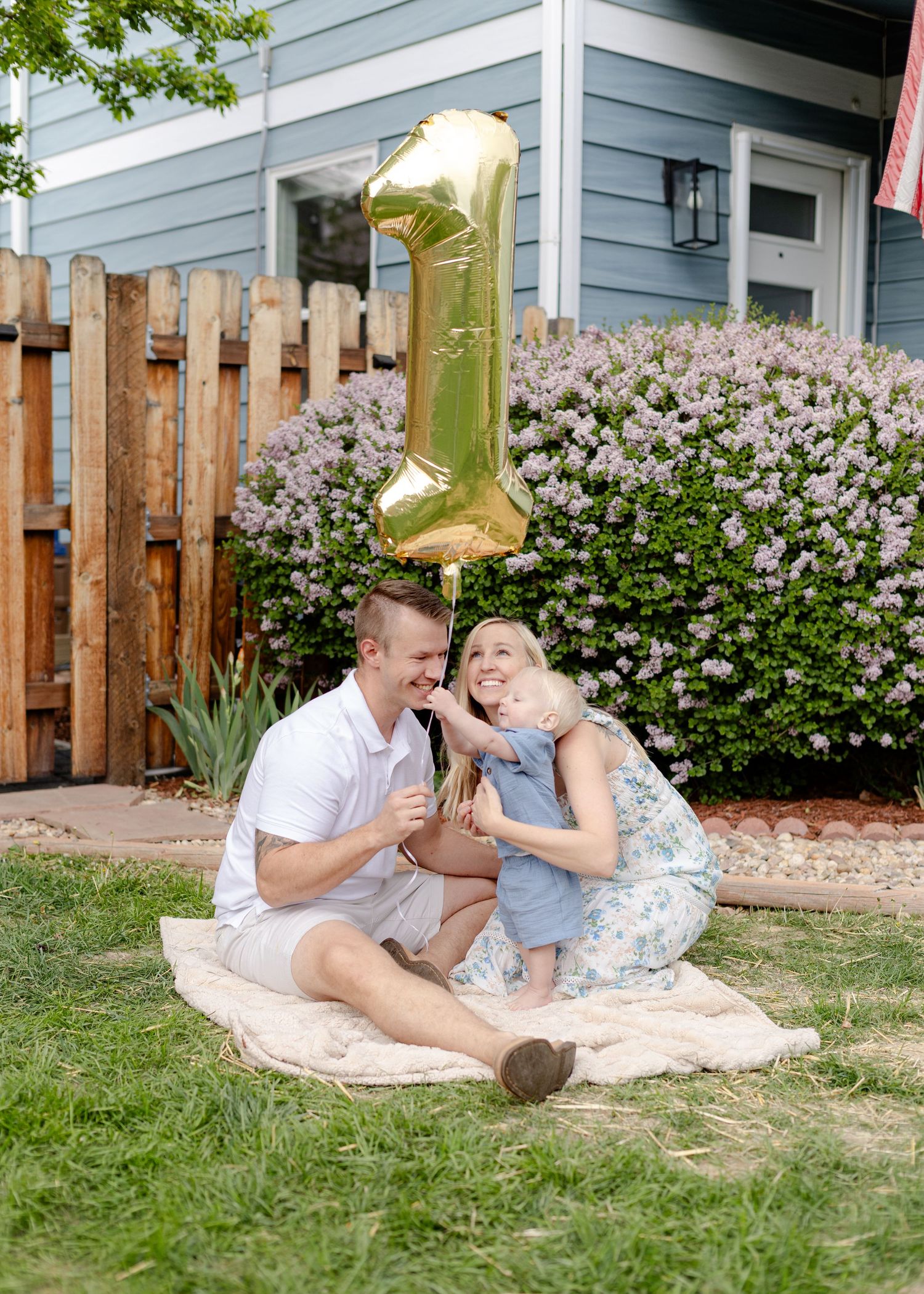Family celebrating baby's first birthday with gold number one balloon in backyard garden.