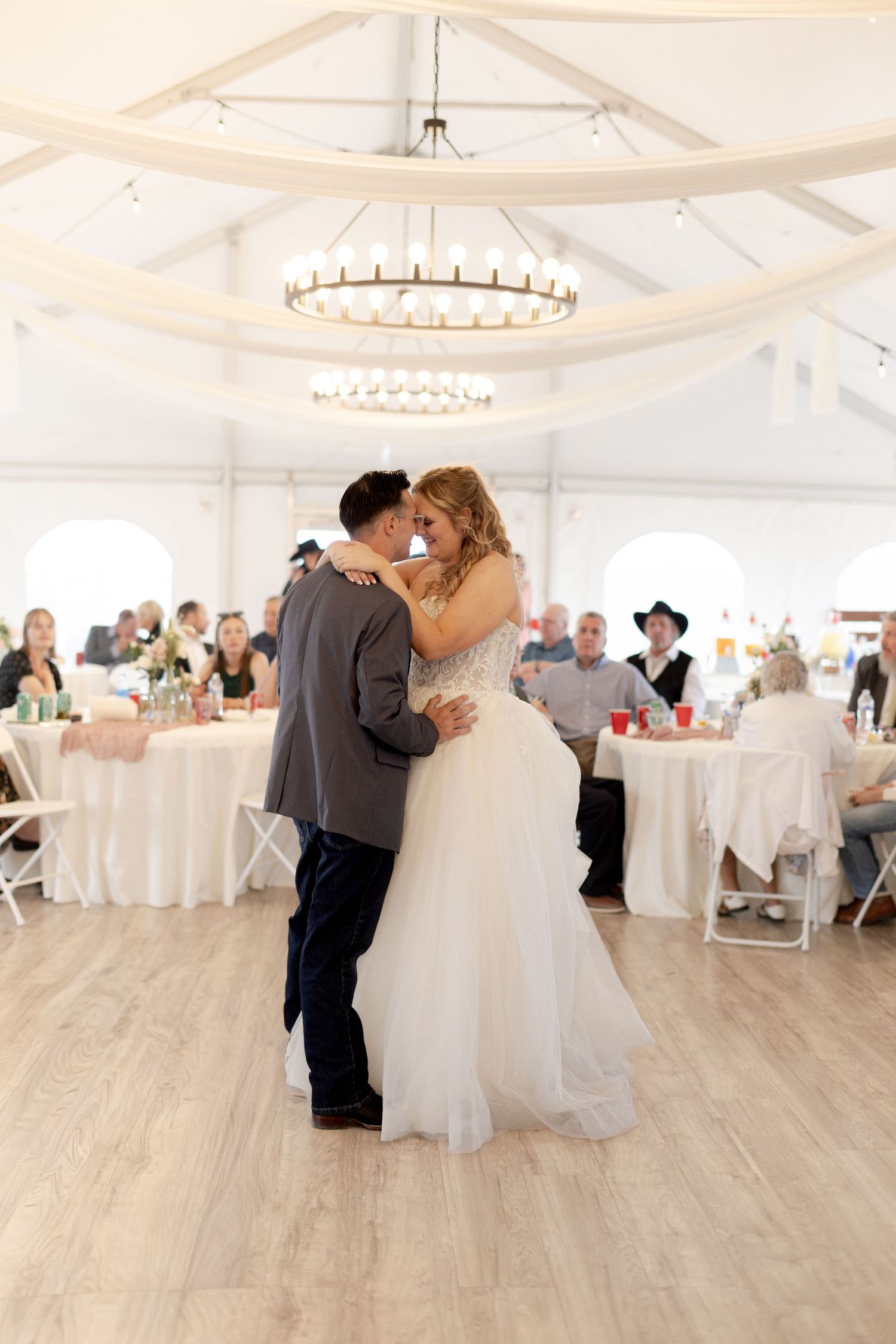 Bride and groom share their first dance in an elegant white tent reception hall with chandelier lighting.