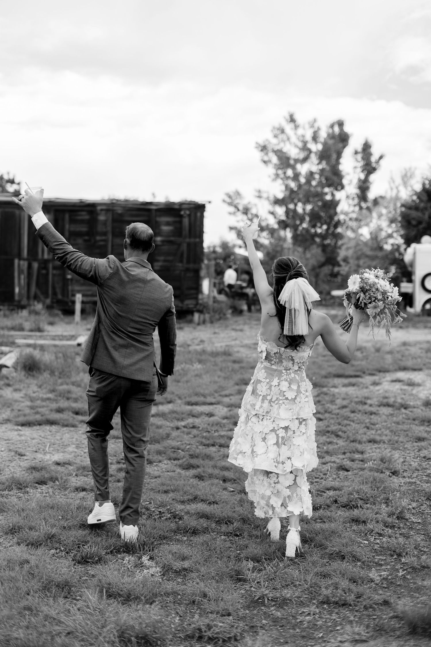 Black and white photo of a bride and groom walking away, arms raised in celebration, bride holding bouquet.