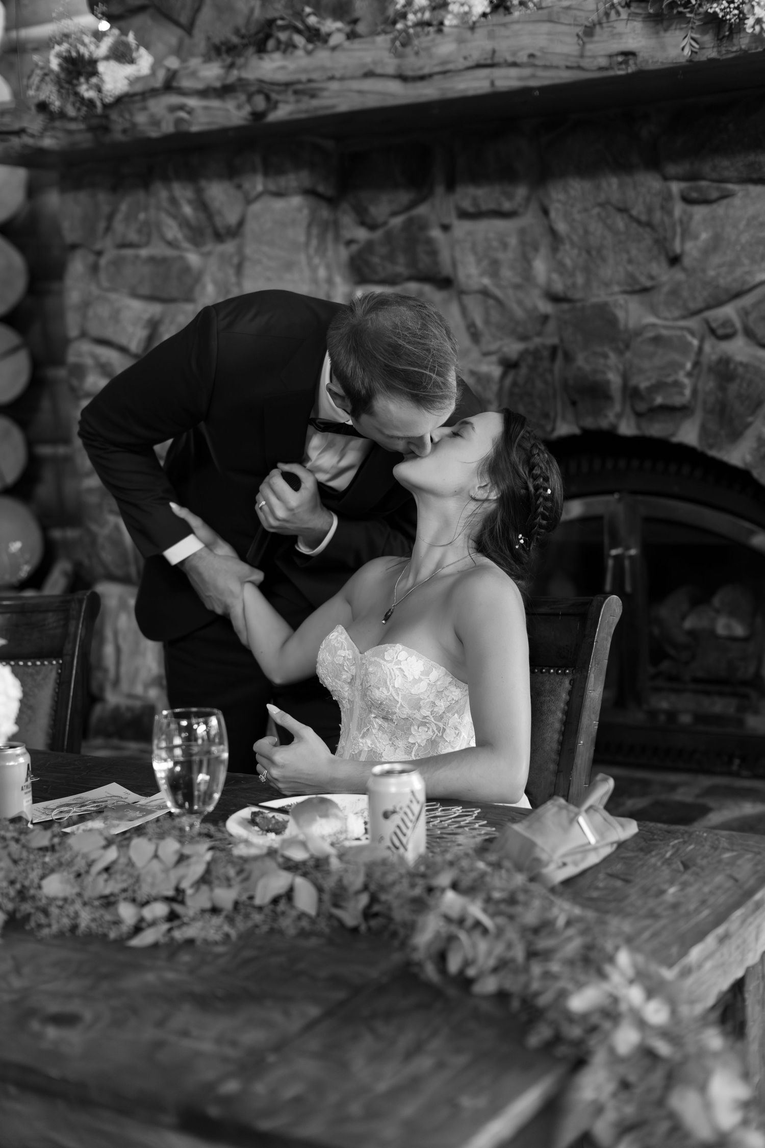 Black and white photo of a bride and groom sharing a kiss at their wedding reception table near a stone fireplace.