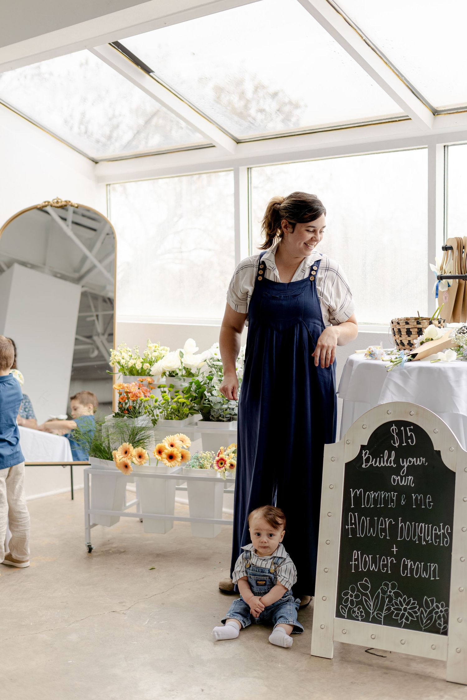 Woman in navy overalls stands beside a chalkboard sign offering $15 DIY flower bouquets and crowns at a bright floral market.