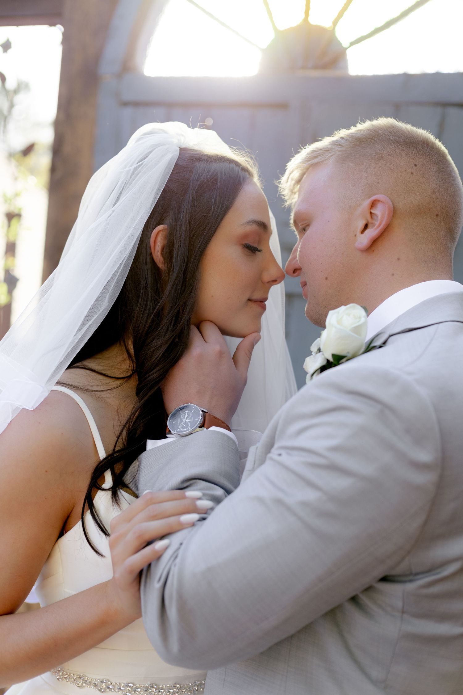 Bride and groom share an intimate moment, nearly kissing, with soft sunlight glowing behind them at their wedding.